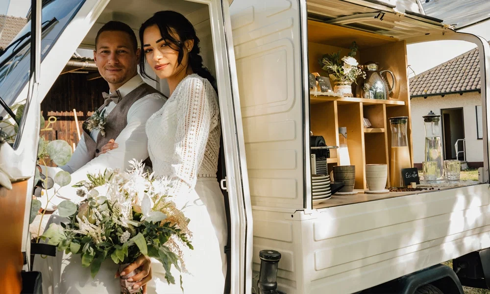 Das Brautpaar posiert in einem weißen mobilen Café-Wagen mit romantischen Boho-Akzenten, einem Blumenstrauß und stilvollen Getränkestation-Details.