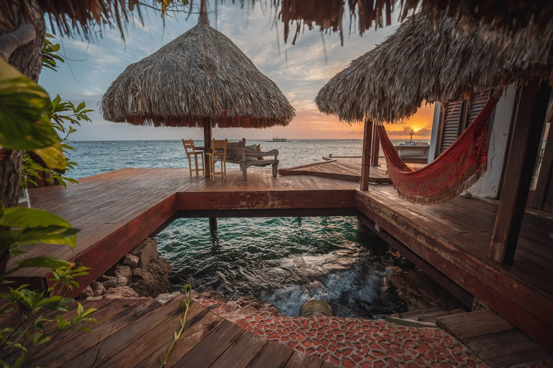 Eine tropische Terrasse mit traditionellem Strohdach bietet einen malerischen Blick auf das Meer bei Sonnenuntergang. Eine Hängematte und Holzmöbel schaffen eine gemütliche Atmosphäre auf der Terrasse, während die Wellen sanft an die Felsen schlagen.