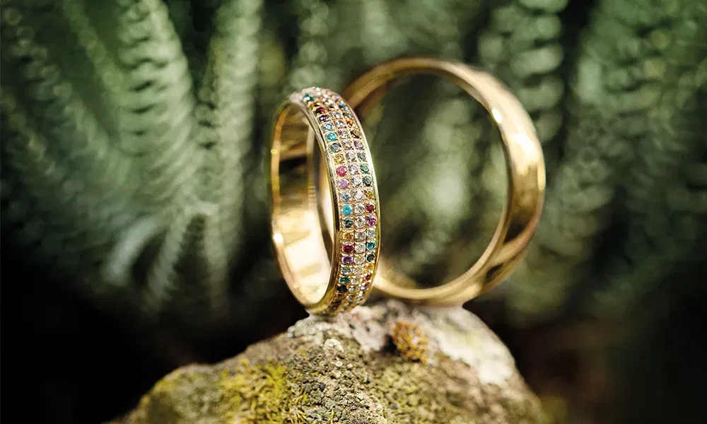 Nahaufnahme zweier goldener Trauringe, einer mit bunten Edelsteinen in einem stilvollen Muster besetzt, arrangiert auf einem Stein mit einem grünen, unscharfen Naturhintergrund.