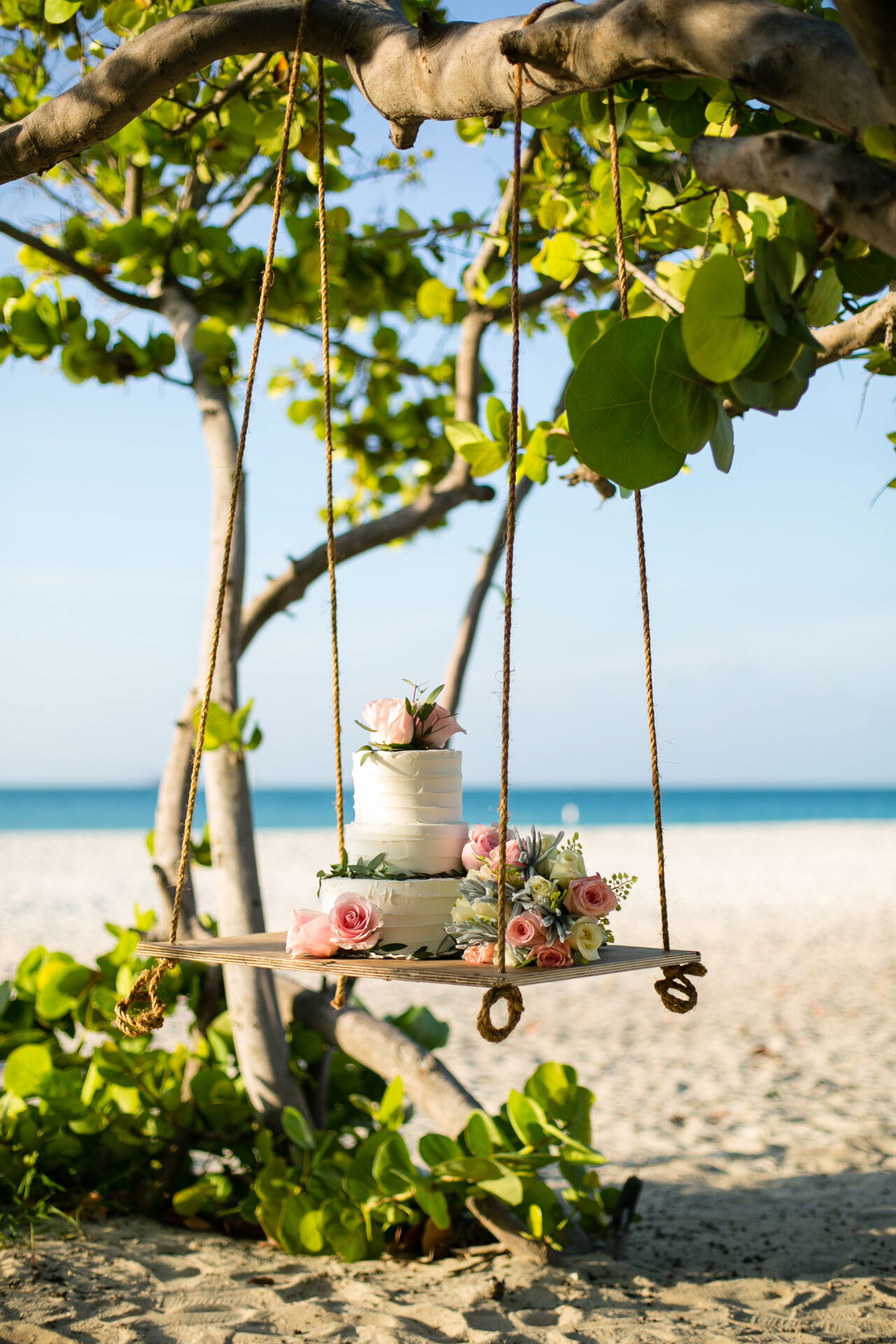 Eine Hochzeitstorte, dekoriert mit Rosen und Grün, steht auf einer hängenden Schaukel aus Holz. Diese ist an einem Baum befestigt und schwebt über dem Sandstrand. Im Hintergrund erstreckt sich das ruhige, blaue Meer unter einem wolkenlosen Himmel.