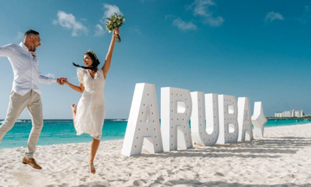 Ein Paar tanzt ausgelassen am Strand von Aruba. Die Braut hält einen Blumenstrauß hoch und trägt ein weißes, knielanges Kleid. Der Bräutigam hält ihre Hand. Im Hintergrund befindet sich ein großes „ARUBA“-Schild, der Strand ist hell, und das Meer leuchtet in türkisblauen Tönen.