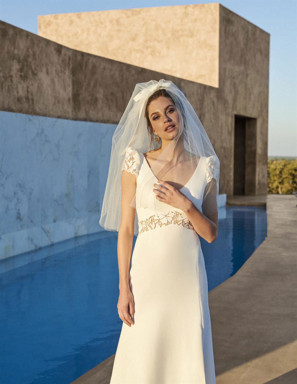 Eine Braut, gekleidet in ein schlichtes, weißes Hochzeitskleid mit zarten Spitzendetails an den Ärmeln und der Taille, steht vor einem Pool unter hellem Sonnenlicht. Sie trägt einen langen, durchsichtigen Schleier, der vom Wind bewegt wird. Hinter ihr erstreckt sich ein modernes, minimalistisches Gebäude aus beigefarbenem Stein, das den zeitgenössischen Stil des Fotos unterstreicht.
