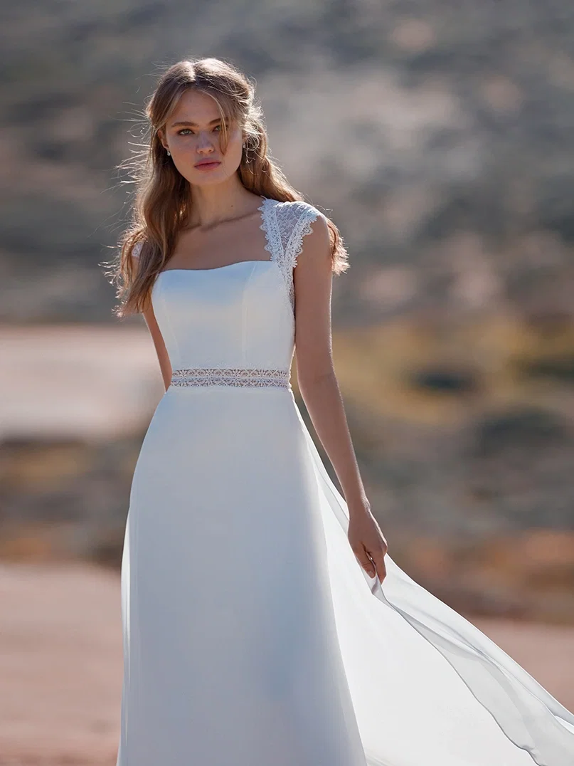 Eine Braut in einem modernen, minimalistischen Brautkleid mit zarten Spitzenakzenten und einem breiten, eckigen Ausschnitt steht vor einer trockenen, hügeligen Kulisse. Der Rock des Kleides weht leicht im Wind, und ihr blondes Haar fällt in sanften Wellen über ihre Schultern. Die warme, trockene Umgebung verstärkt den Kontrast zum weichen, fließenden Stoff ihres Kleides.