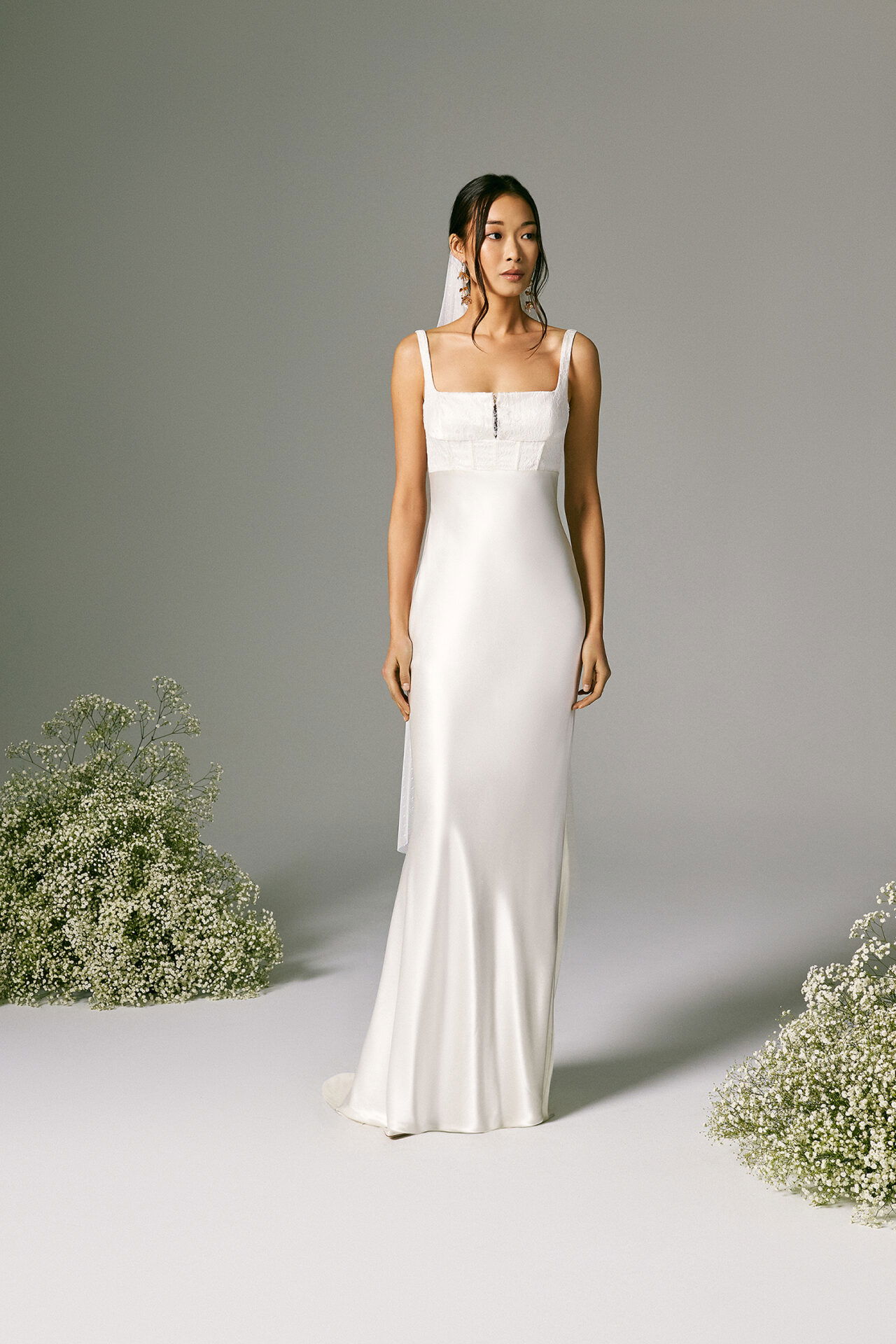 Eine Braut, gekleidet in ein minimalistisches, figurbetontes Brautkleid mit einem geraden, eckigen Ausschnitt und zarten Trägern, steht in einem schlichten Studio vor einer grauen Wand. Ihr schlichtes, modernes Hochzeitskleid wird durch feine Spitzendetails am Oberteil akzentuiert. Um sie herum sind Büschel von Schleierkraut drapiert, was eine natürliche und beruhigende Kulisse schafft.