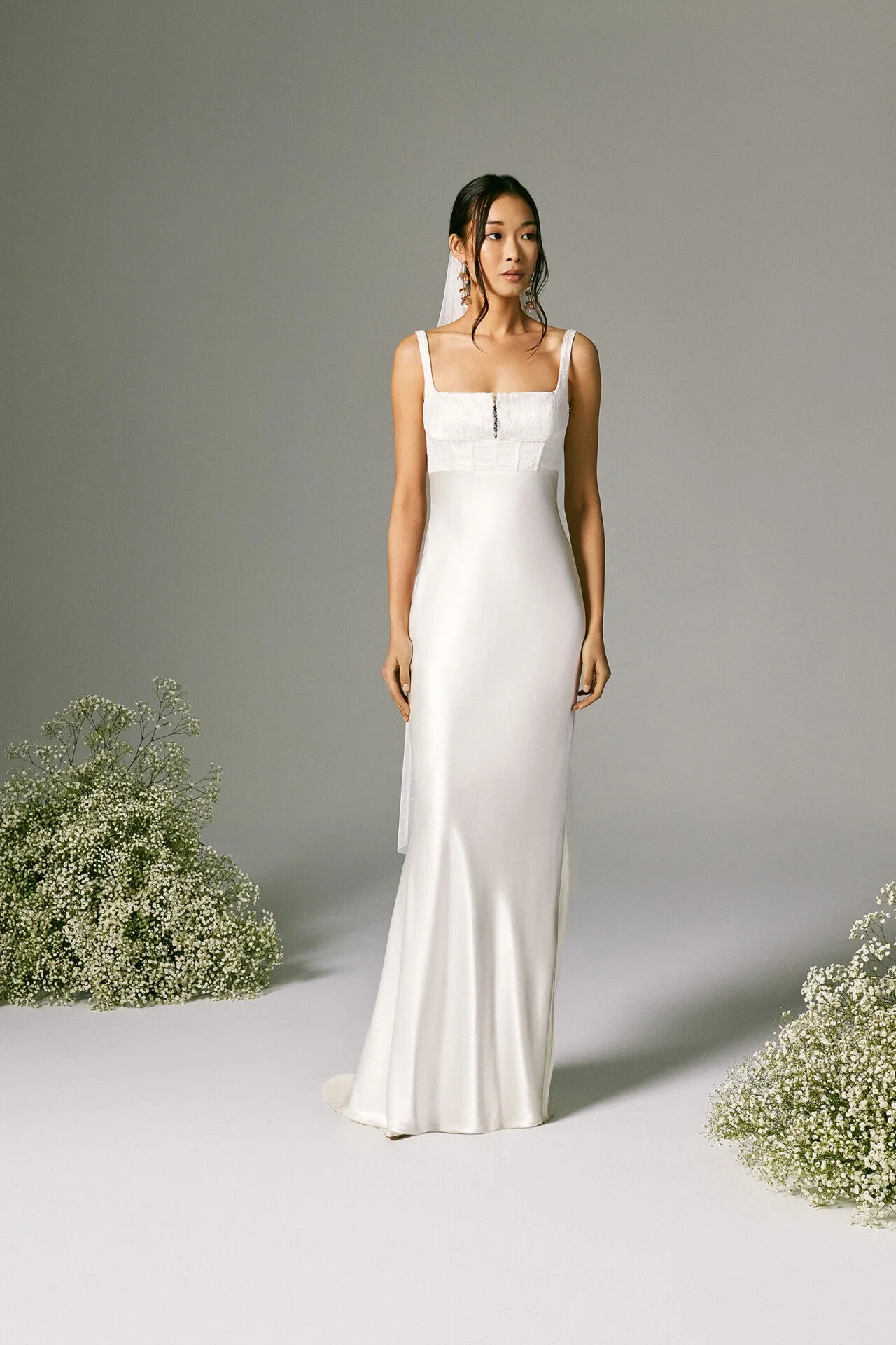 Eine Braut, gekleidet in ein minimalistisches, figurbetontes Brautkleid mit einem geraden, eckigen Ausschnitt und zarten Trägern, steht in einem schlichten Studio vor einer grauen Wand. Ihr schlichtes, modernes Hochzeitskleid wird durch feine Spitzendetails am Oberteil akzentuiert. Um sie herum sind Büschel von Schleierkraut drapiert, was eine natürliche und beruhigende Kulisse schafft.