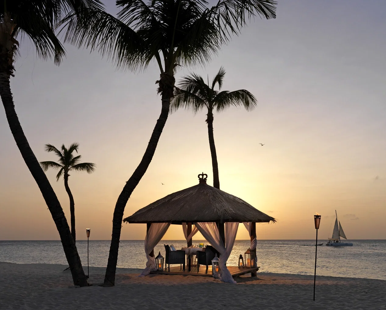 Ein Pavillon aus Holz und Strohdach ist am Strand bei Sonnenuntergang für ein romantisches Dinner arrangiert. Der Tisch ist für zwei Personen gedeckt, und Fackeln beleuchten den Weg. Palmen und das ruhige Meer schaffen eine idyllische Szenerie.
