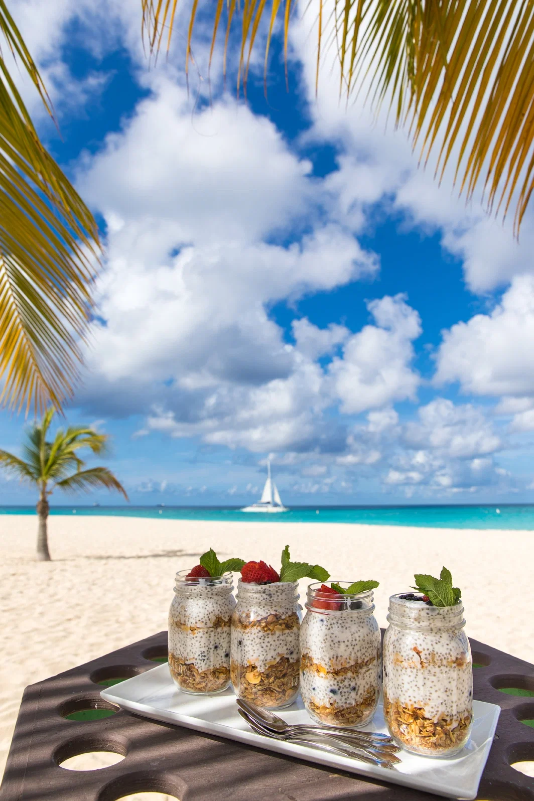 Eine Auswahl an Chia-Pudding-Gläsern mit Beeren und Minze steht am Strand auf einem Tablett. Palmenblätter rahmen die Szene ein, während das Meer und ein Segelboot im Hintergrund unter einem sonnigen Himmel zu sehen sind.