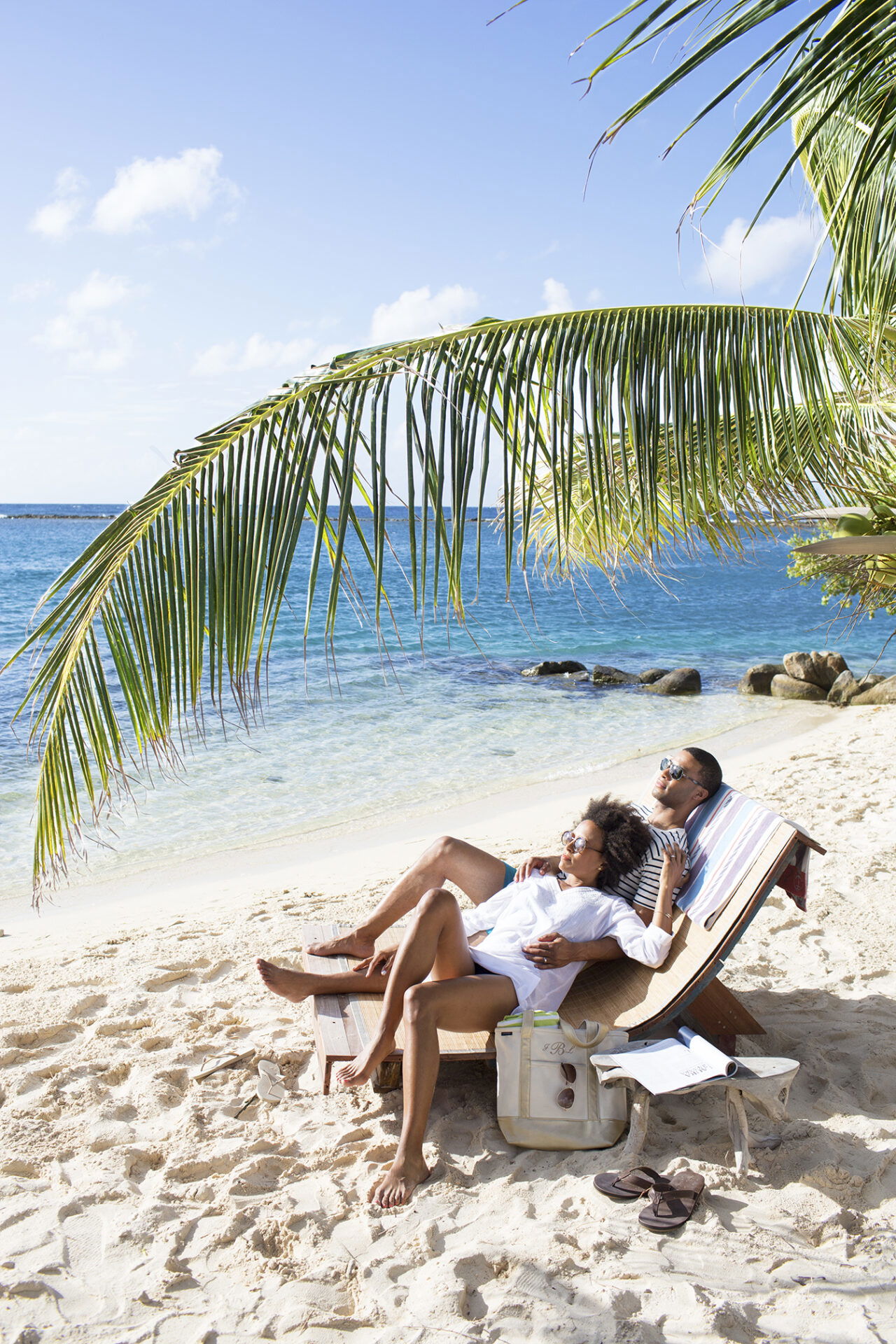Unter dem Schatten einer Palme entspannt ein Paar auf einer Liege am Strand. Ihre Füße ruhen im weichen Sand, und das türkisfarbene Meer erstreckt sich im Hintergrund. Eine Tasche und Sandalen liegen neben der Liege.