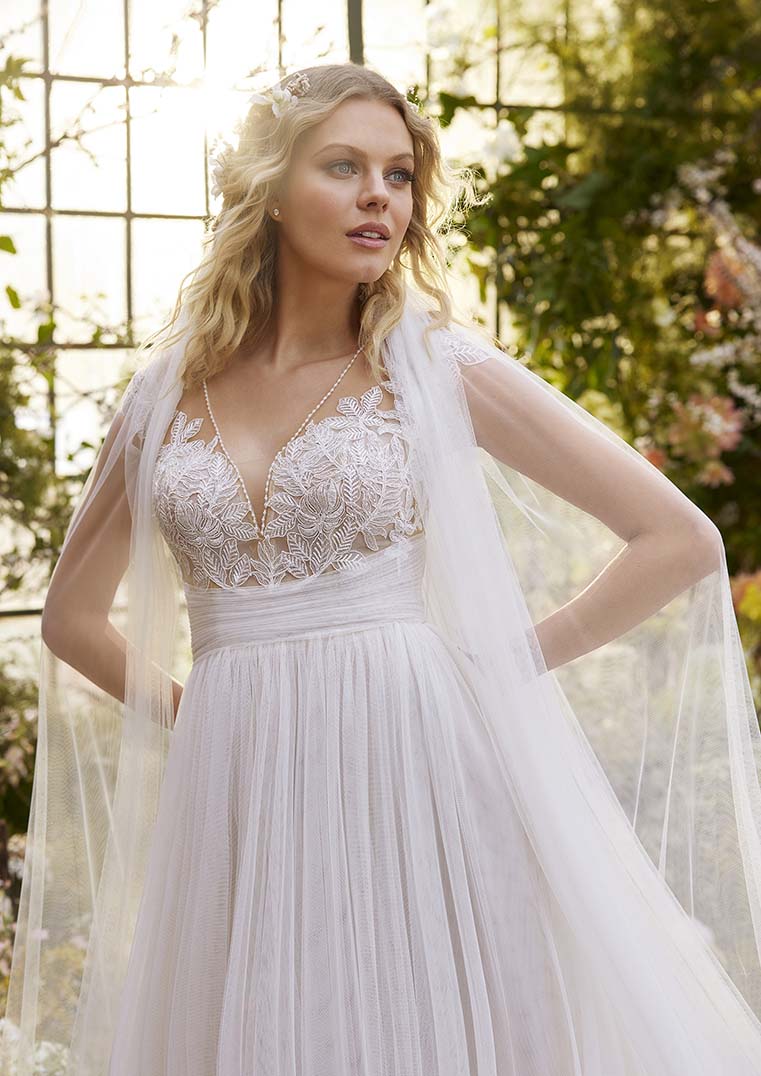 Eine Braut in einem transparenten Tüllkleid mit zarten Blütenmustern steht vor einer verglasten Wand in einem grünen Garten. Der weiche Tüll und die floralen Verzierungen geben dem Kleid einen märchenhaften Charakter.