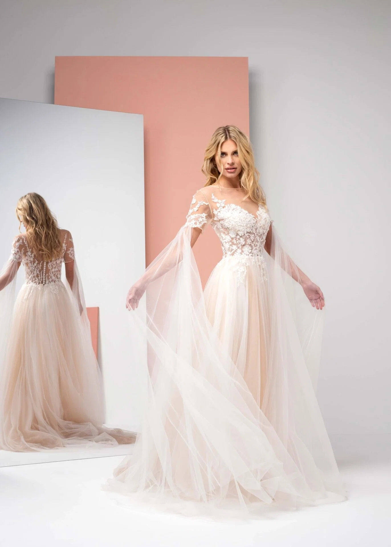 Braut in einem modernen, transparenten Tüllkleid mit floralen Verzierungen und fließenden Ärmeln steht vor einem Spiegel. Das elegante, minimalistische Design der Szene unterstreicht die zeitgemäße und schlichte Eleganz des Kleides.