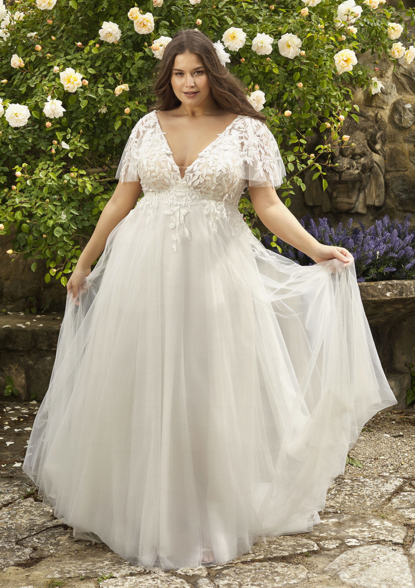 Braut in einem fließenden Tüllkleid mit zarten Ärmeln und eleganter Spitze, umgeben von Blumen.