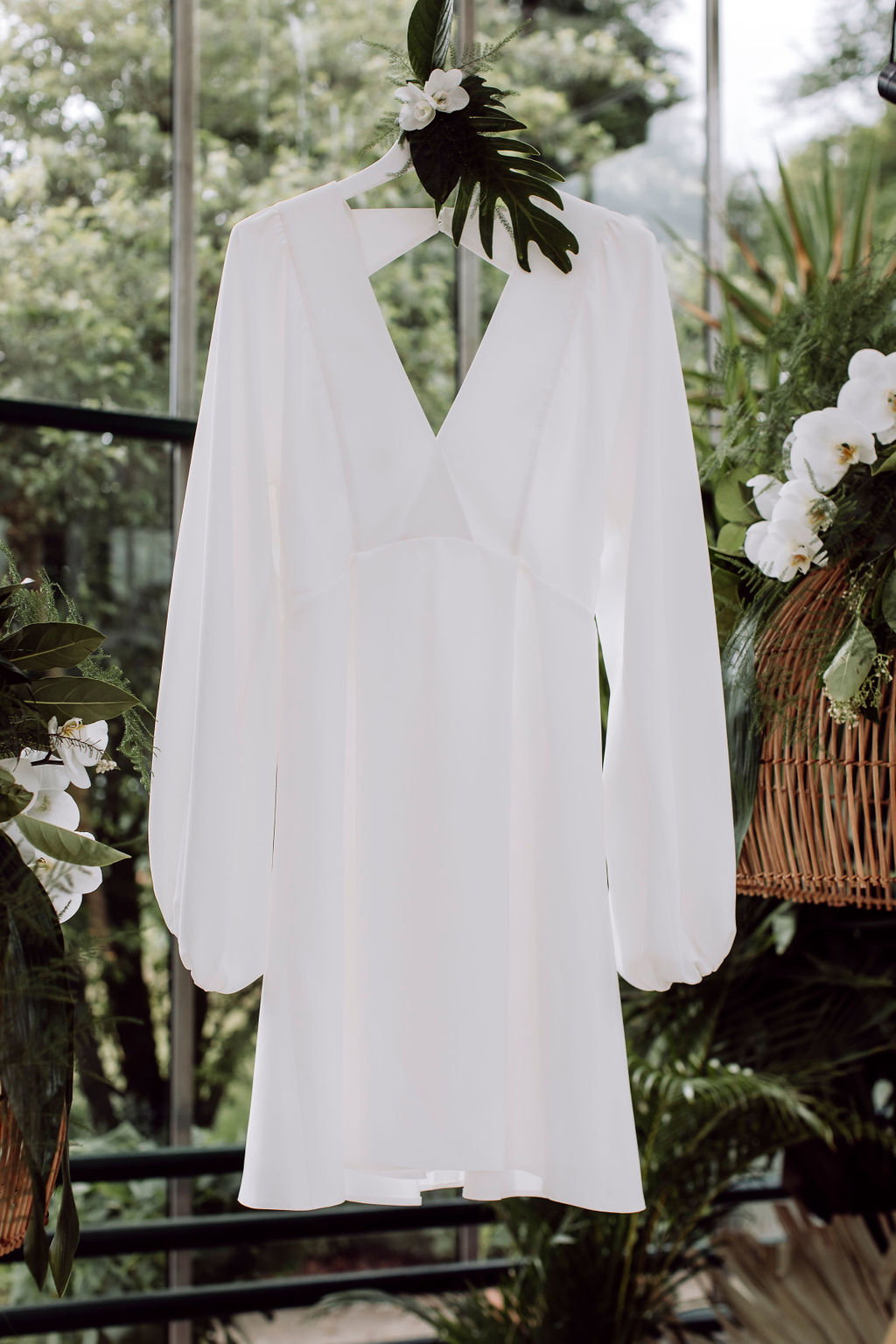 Weißes Brautkleid in einem Gewächshaus, umgeben von exotischen Pflanzen wie Orchideen und Monstera-Blättern für tropische Hochzeit.