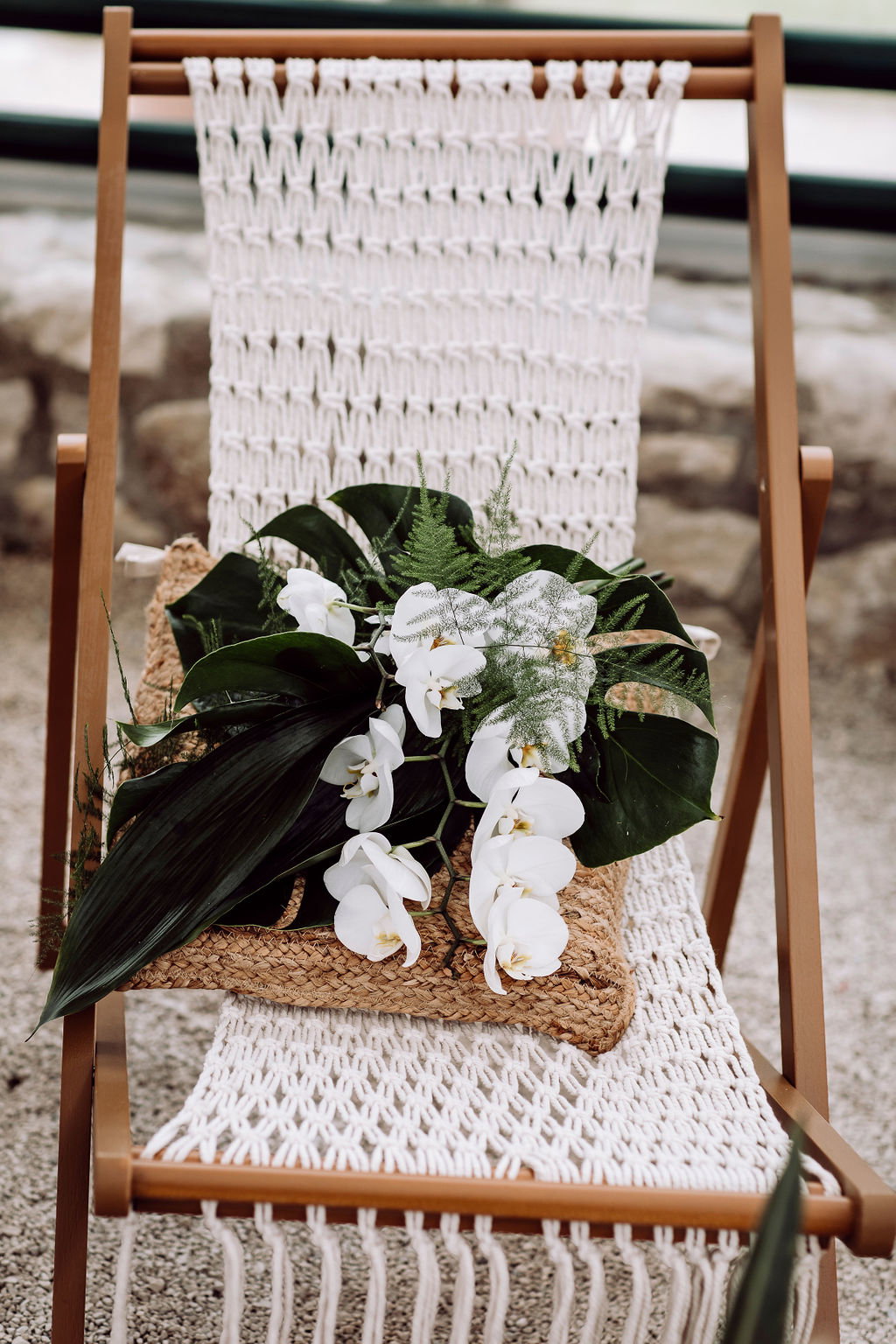 Tropisch inspirierter Brautstrauß aus Orchideen und Monstera-Blättern, dekorativ platziert auf einem Makramee-Stuhl bei einer Boho-Hochzeit.