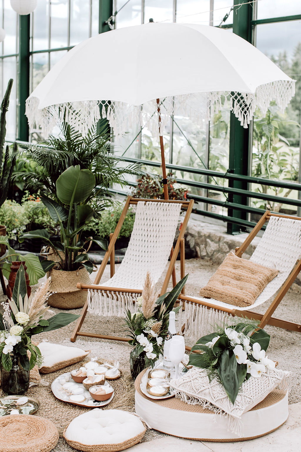 Entspannte Hochzeitsecke mit Liegestühlen, Palmen, Bodenkissen und einem Makramee-Sonnenschirm, ideal für eine tropische Feier.