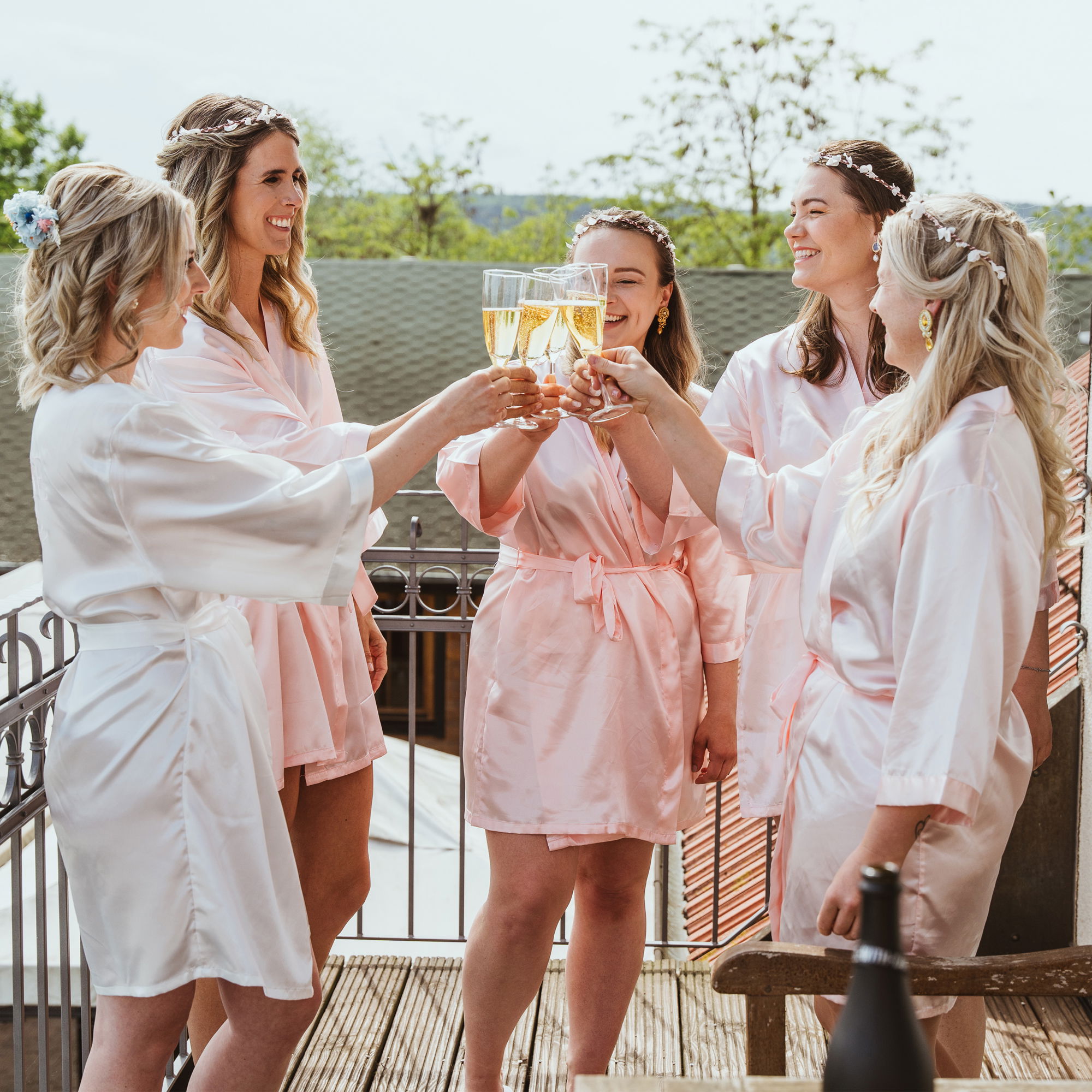 Braut und ihre Brautjungfern feiern im Freien, tragen Seidenroben und stoßen mit Champagner an, während sie sich auf die Hochzeit vorbereiten.