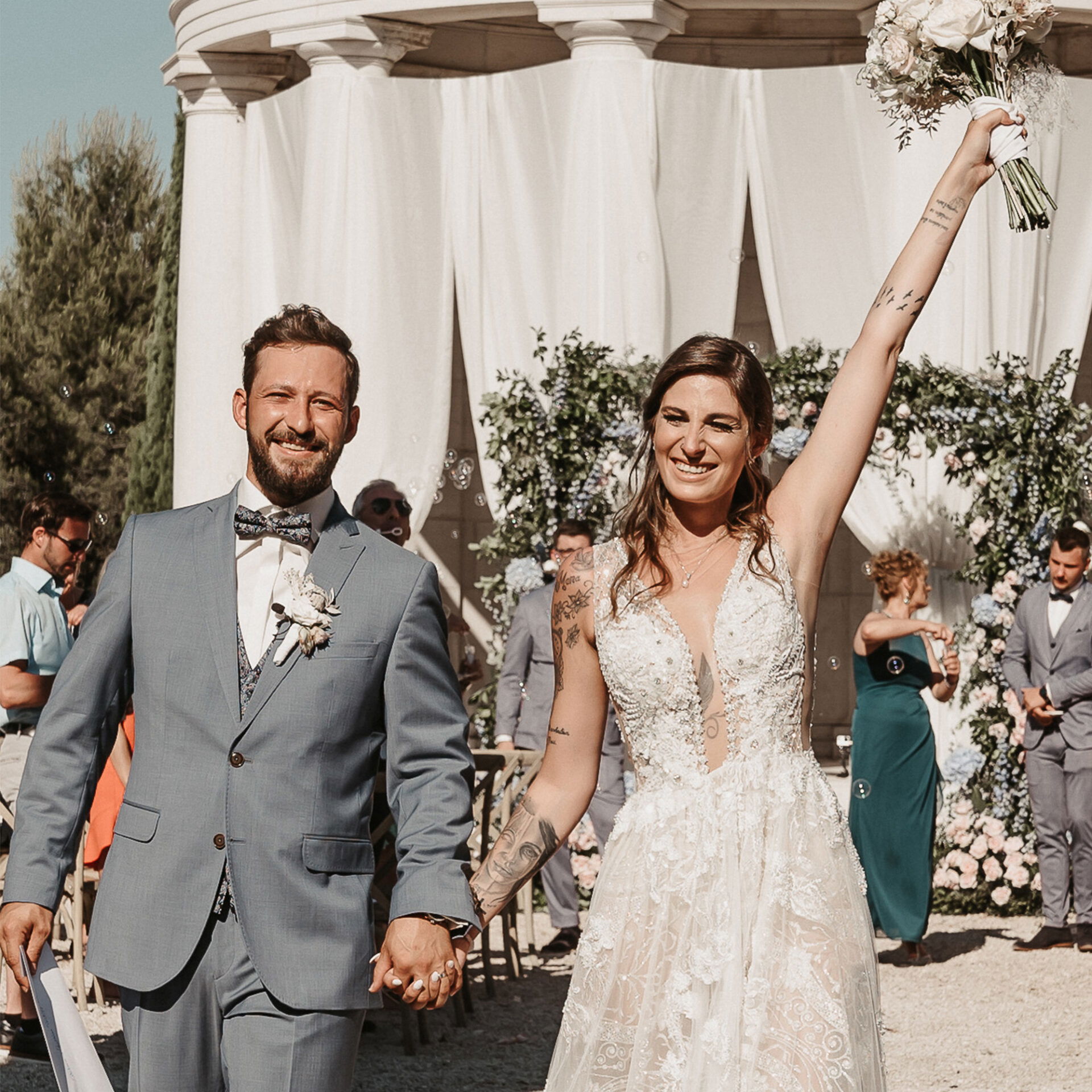 Fröhliches Brautpaar nach der Trauung, die Braut jubelt mit erhobenem Brautstrauß, während sie mit dem Bräutigam den Auszug genießt.