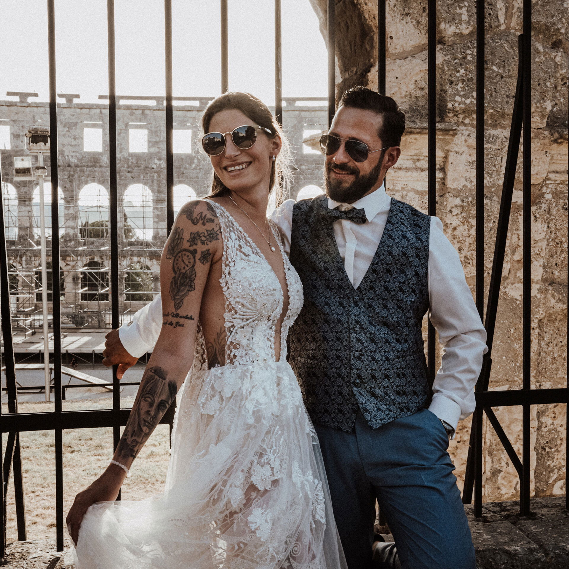 Lächelndes Brautpaar in Sonnenbrillen, elegant gekleidet, posiert vor einem historischen Monument an ihrem Hochzeitstag.