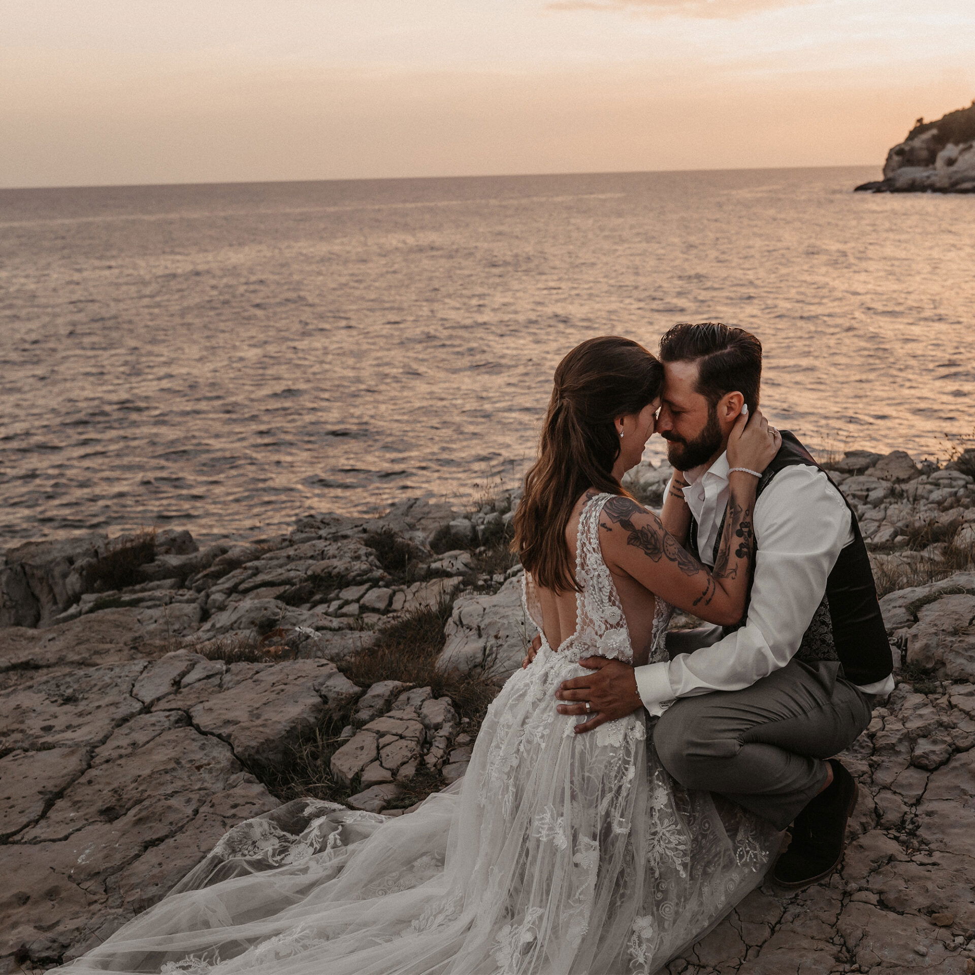 Brautpaar sitzt liebevoll umarmt am Meer, das Brautkleid fließt über die Felsen, während die Sonne langsam hinter dem Horizont verschwindet.