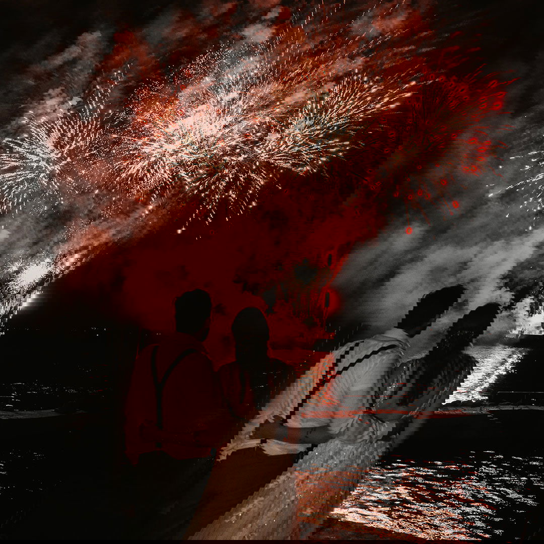 Braut und Bräutigam blicken auf ein eindrucksvolles Feuerwerk am Nachthimmel, das über dem Wasser explodiert und den Hochzeitstag krönt.