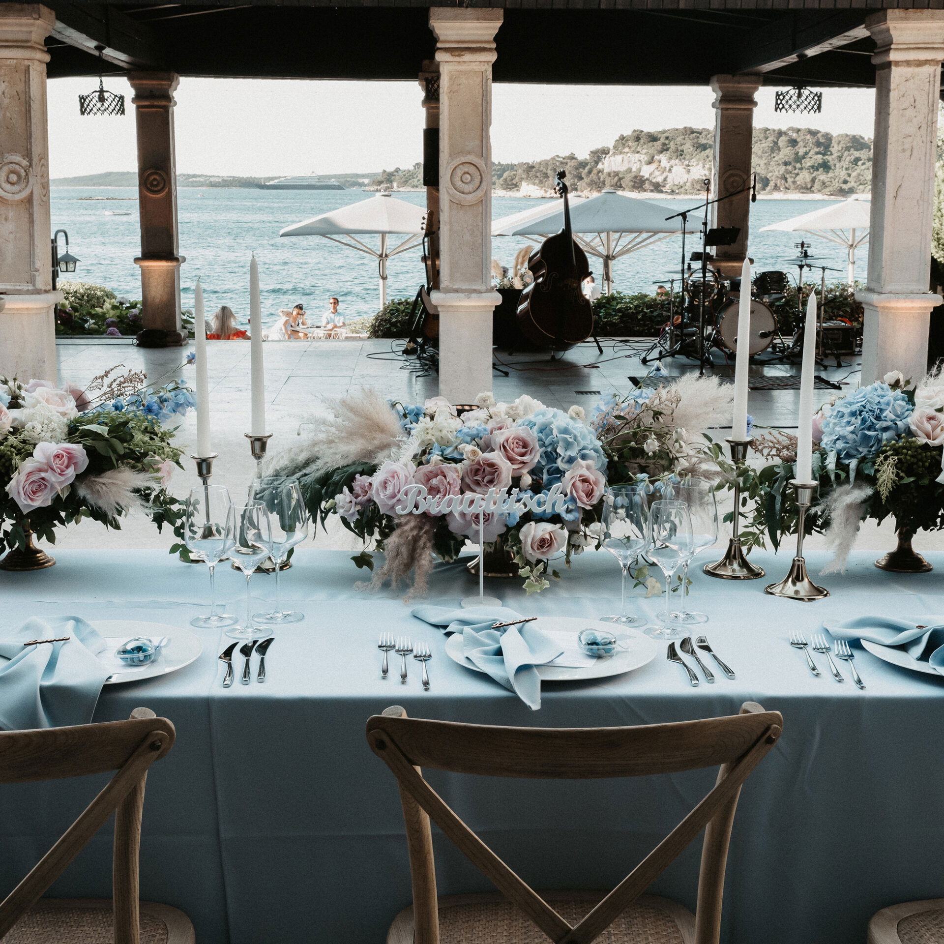 Hochzeitstafel mit aufwendiger Blumendekoration in Blau und Rosa, weißen Kerzen und Meerblick im Hintergrund, bereit für den Empfang.