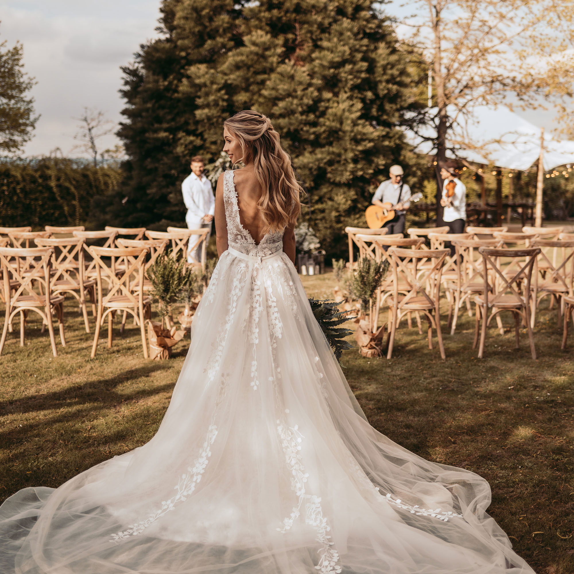 Die Braut, gekleidet in ein atemberaubendes weißes Kleid, nähert sich den aufgestellten Stühlen der Hochzeitsgesellschaft unter freiem Himmel.