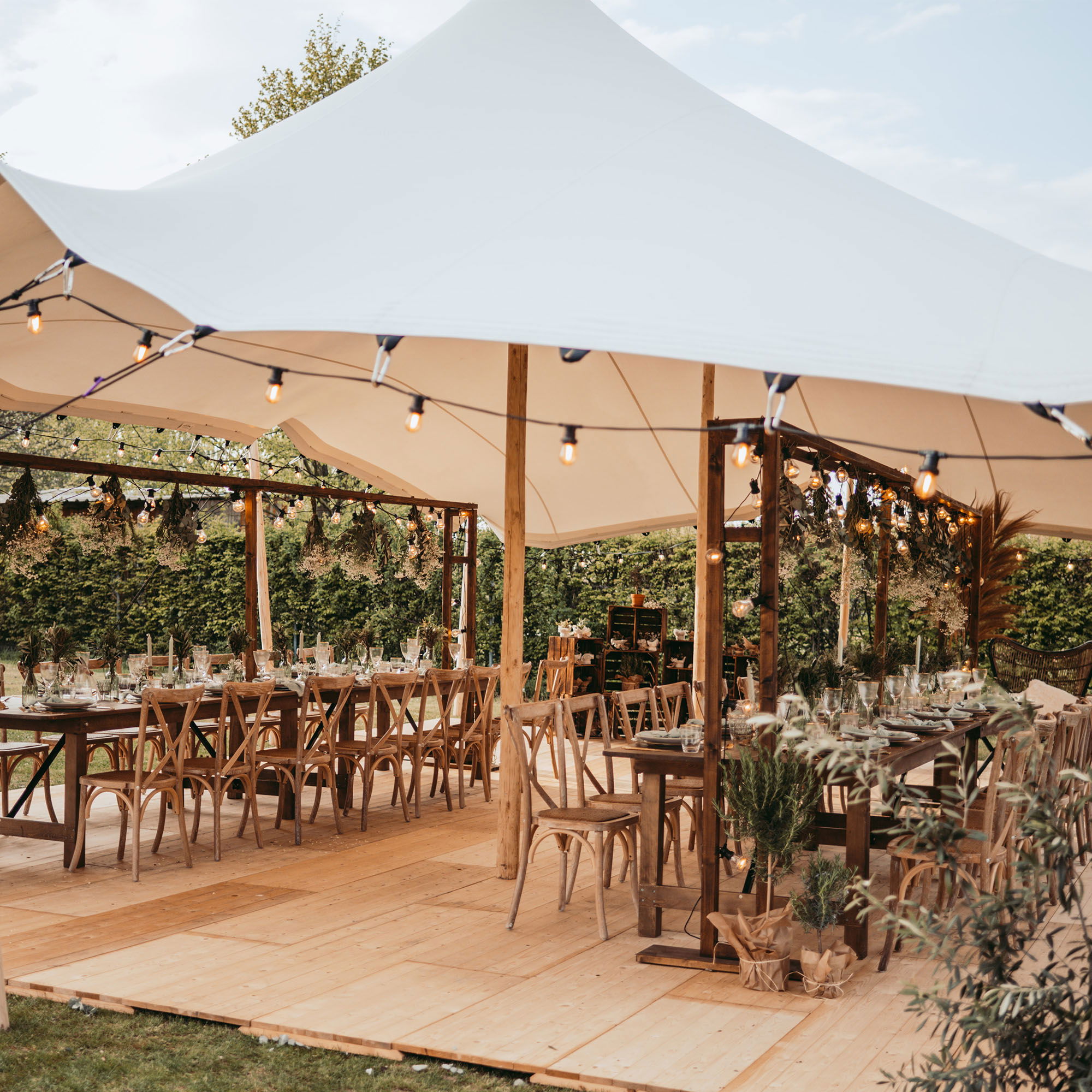 Große Outdoor-Hochzeitslocation unter einem dekorativen Zelt mit festlich gedeckten Tischen, Holzboden und gemütlichen Lichterketten.