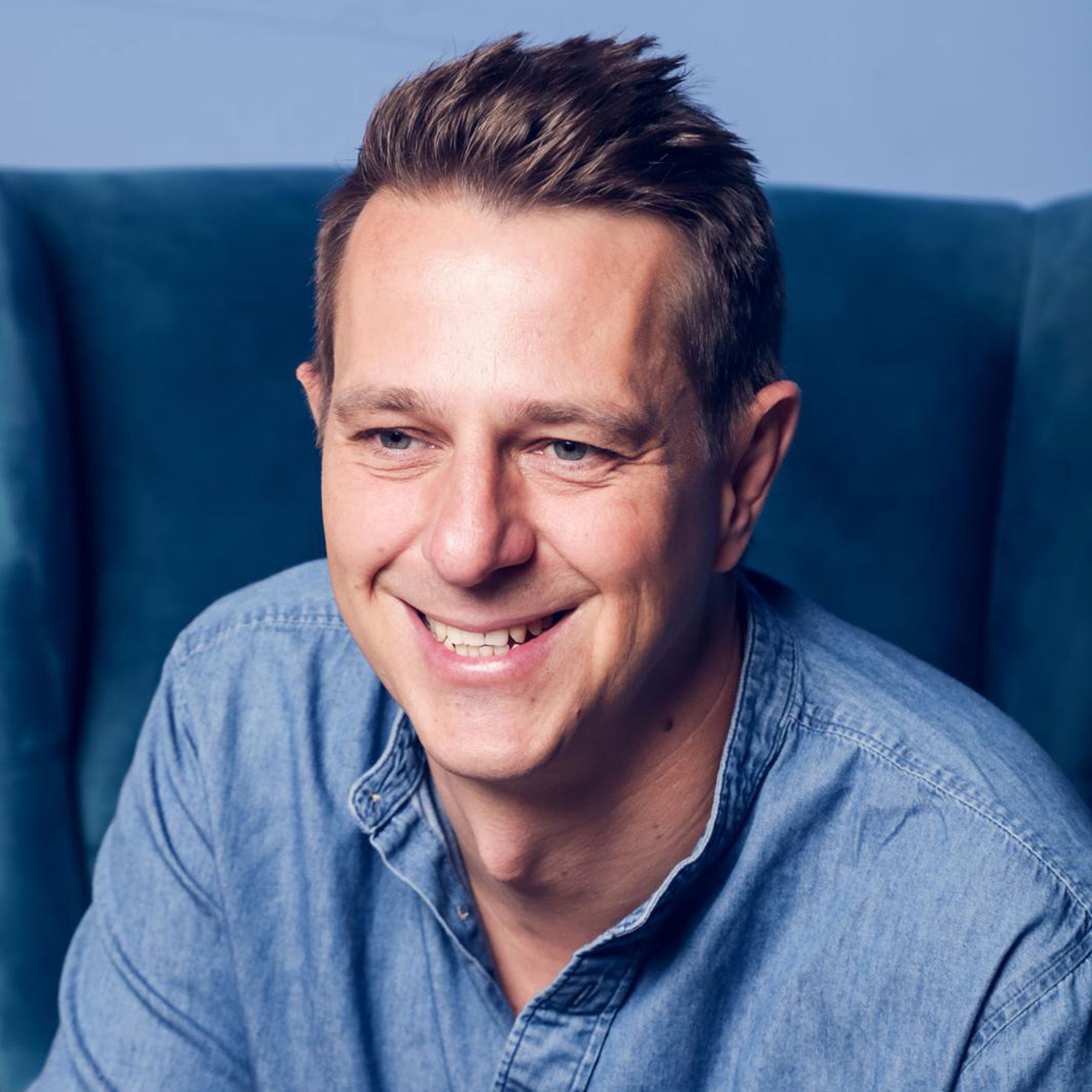 Ein lächelnder Mann mit kurzen braunen Haaren trägt ein lässiges, blaues Hemd und sitzt auf einem blauen Stuhl. Der Hintergrund ist ebenfalls in Blautönen gehalten.