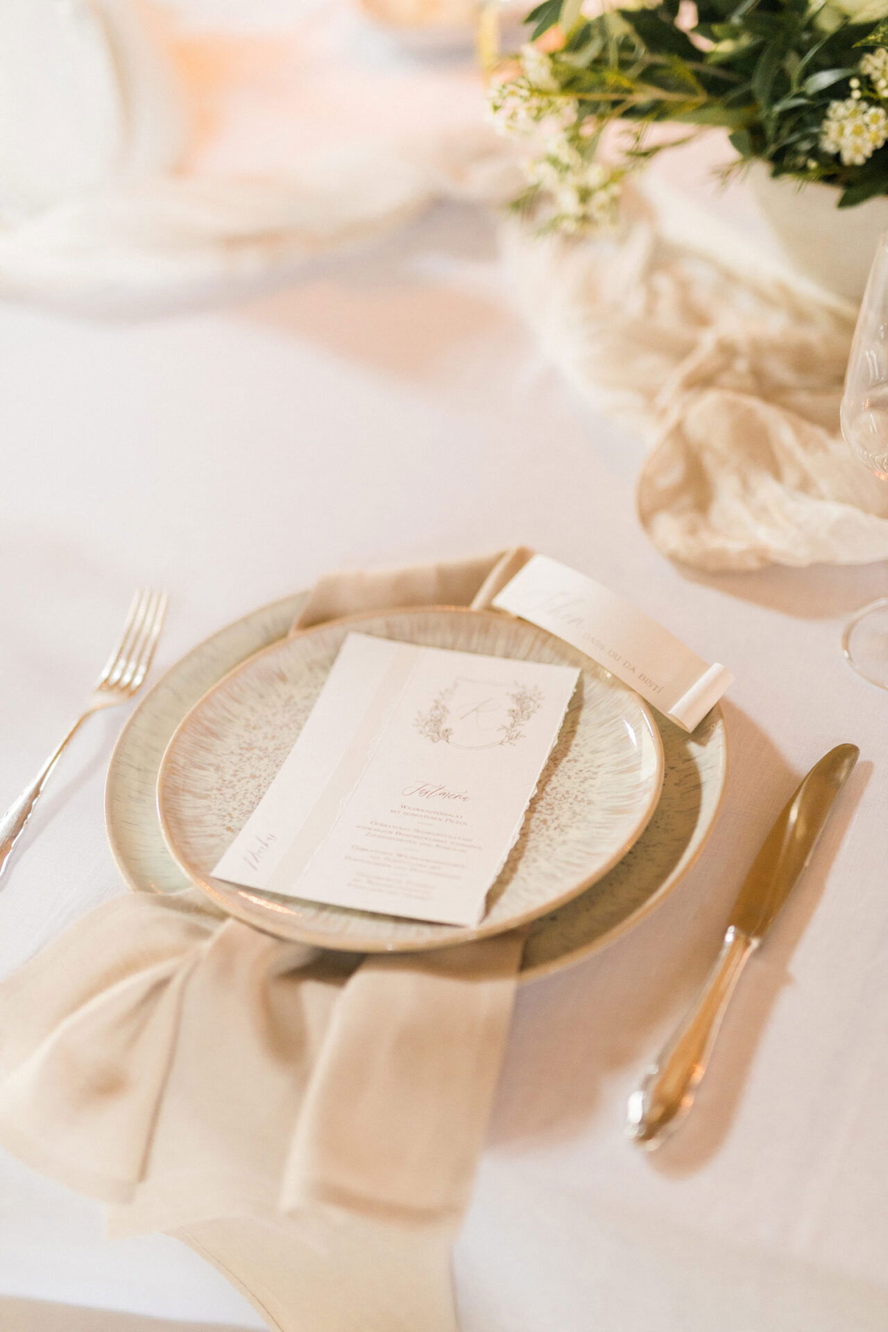 Detailaufnahme einer Hochzeitstafel mit weißem Teller, einer stilvoll gefalteten Stoffserviette und einer Menükarte, die auf dem Gedeck liegt.
