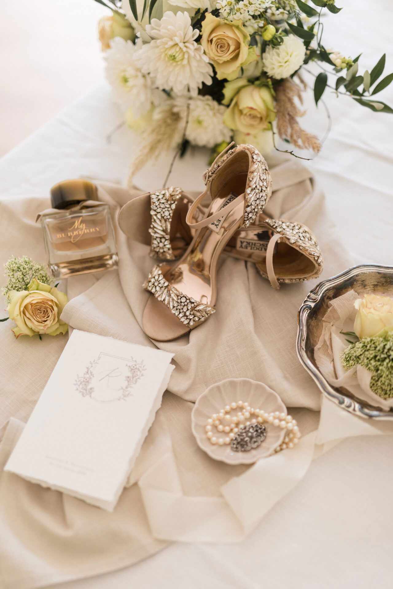 Ein elegant gestaltetes Stillleben einer Hochzeit mit funkelnden, beigen Sandaletten, die mit Schmucksteinen verziert sind, einem Glasflakon mit Parfüm, einer weißen Einladungskarte und einem Perlenarmband, das in einer kleinen Schale auf Leinenstoff liegt. Im Hintergrund ist ein Bouquet aus weißen und cremefarbenen Rosen sowie anderen Blumen zu sehen.