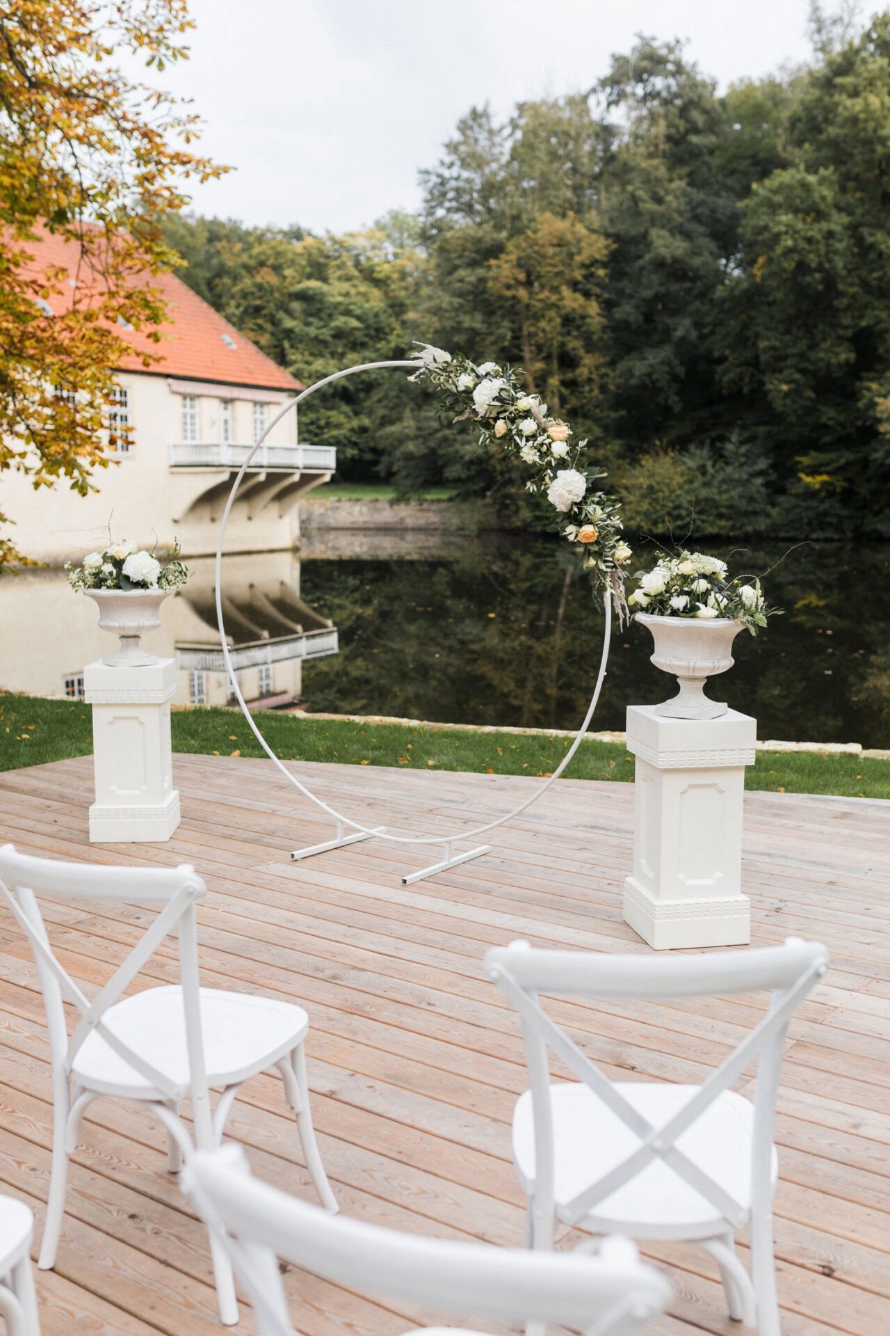 Ein minimalistischer runder Blumenbogen bildet den Mittelpunkt einer Hochzeitszeremonie im Freien. Umgeben von weißen Stühlen und sanft dekorierten Blumensäulen ist die Szene von einem ruhigen Gewässer und herbstlichem Laub eingerahmt.