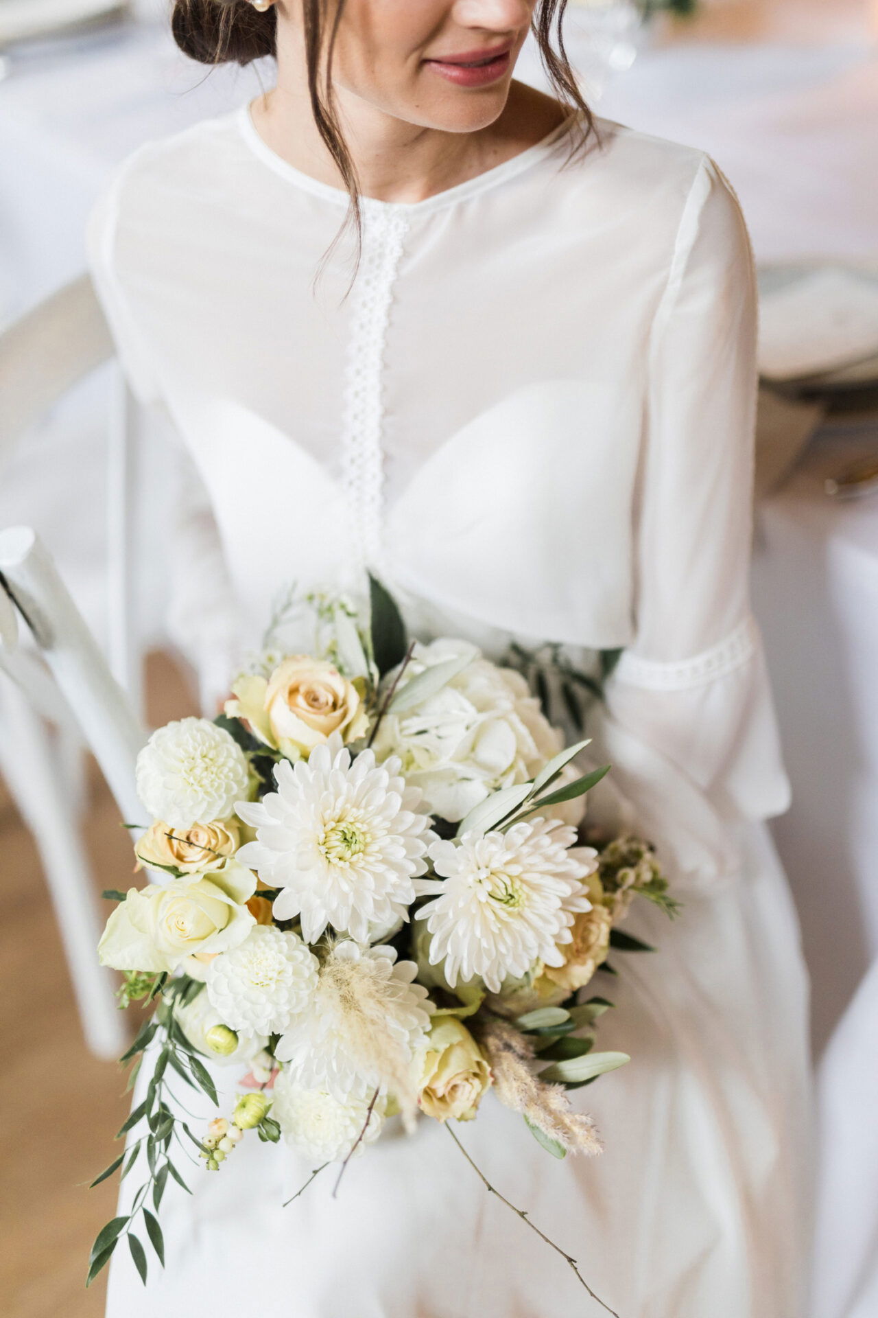 Ein eleganter Brautstrauß mit weißen und cremefarbenen Blumen wird von der Braut in einem zarten, transparenten Kleid mit feinen Details gehalten. Der Strauß besteht aus Dahlien, Rosen und grünen Blättern, die der Szene eine natürliche Note verleihen.