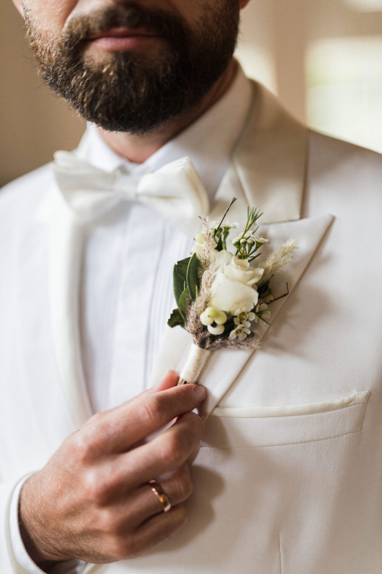 Der Bräutigam in einem klassischen, hellen Anzug richtet seine florale Anstecknadel mit weißen Blumen. Die dezente Kombination aus Rose, Grün und feinen Details ergänzt sein formelles Hochzeitsoutfit auf elegante Weise.