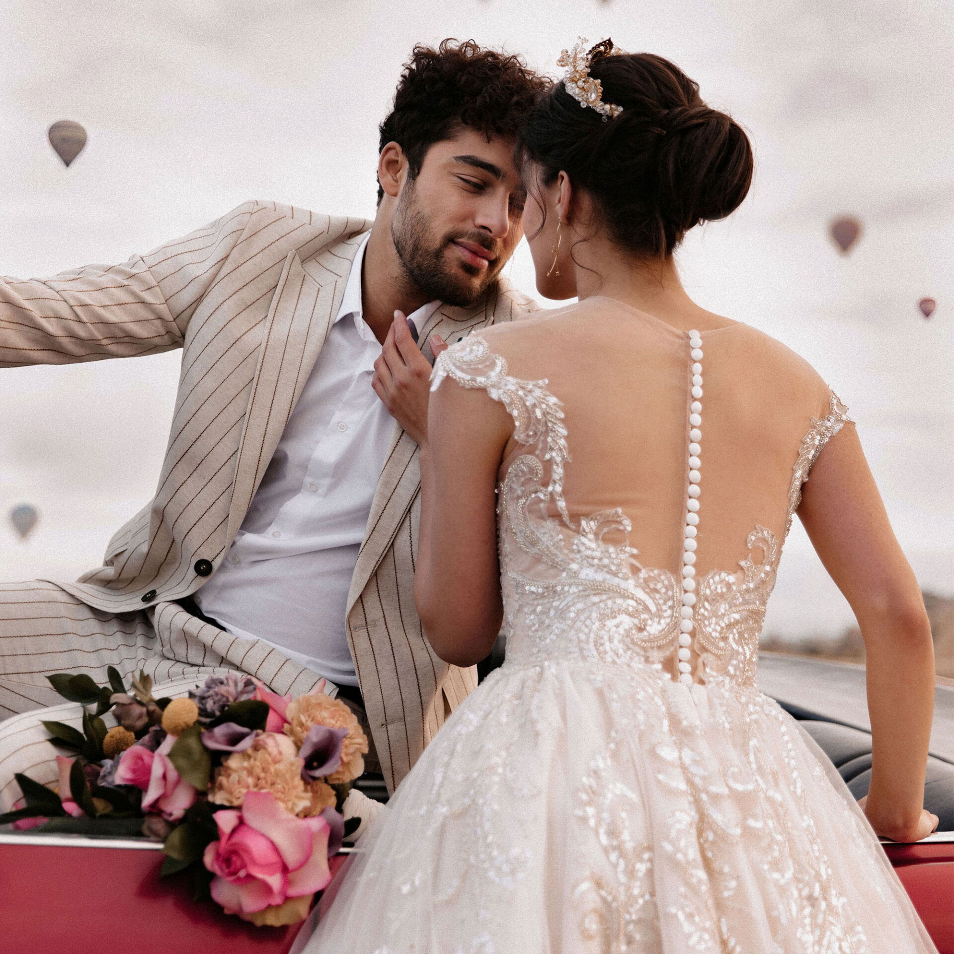 Das Brautpaar lehnt lächelnd an einem Oldtimer-Cabrio, umgeben von einer traumhaften Kulisse mit Heißluftballons.