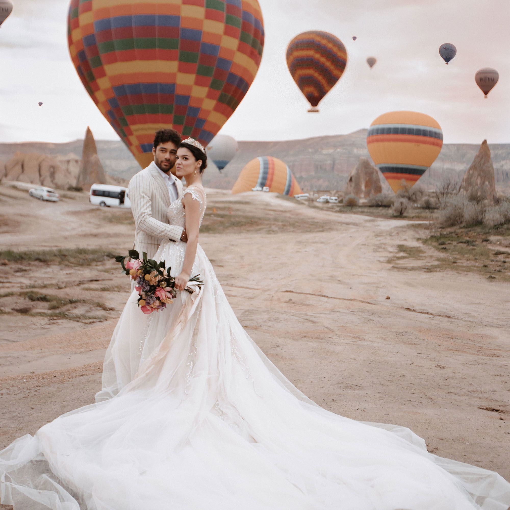 Braut und Bräutigam posieren vor einer atemberaubenden Landschaft mit schwebenden Heißluftballons, die Braut hält einen Brautstrauß.