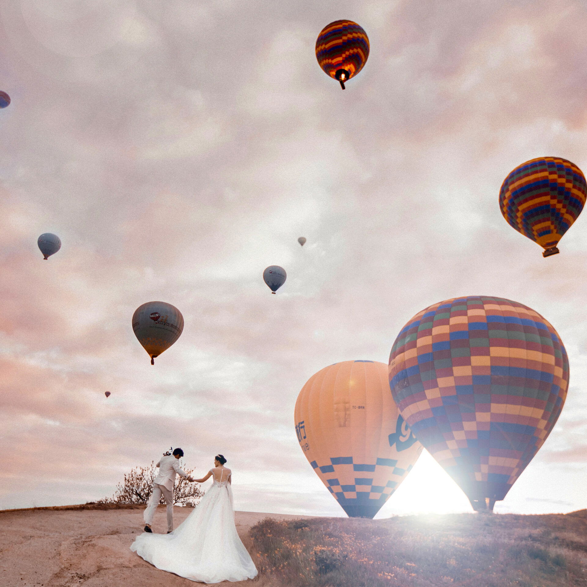 Brautpaar schreitet den Weg entlang, umgeben von Heißluftballons am Himmel, während die Sonne hinter den Hügeln untergeht.