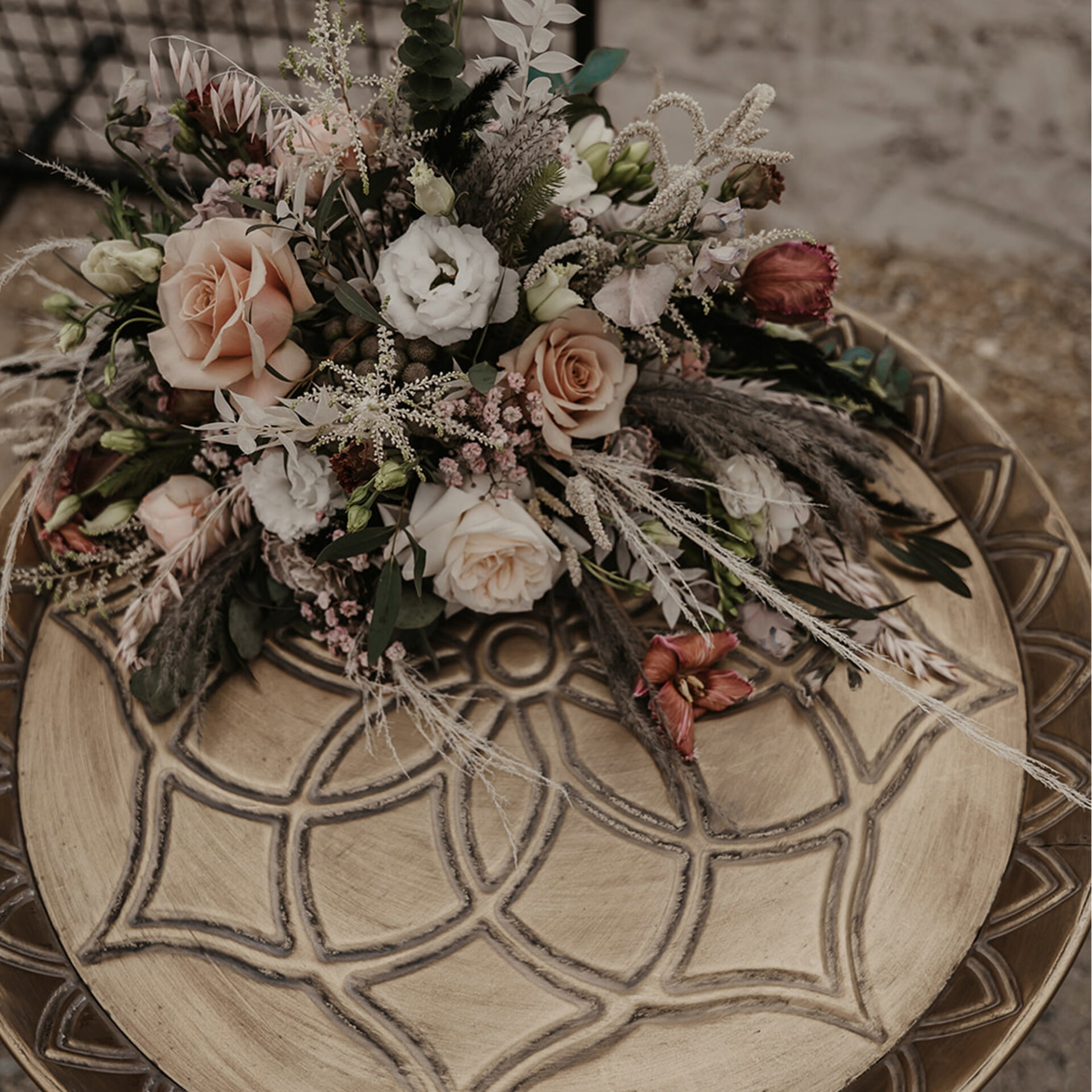 Blumenarrangement mit Rosen und Gräsern auf einem kunstvoll verzierten Holztisch, das durch seine Mischung aus Eleganz und Wildheit besticht.