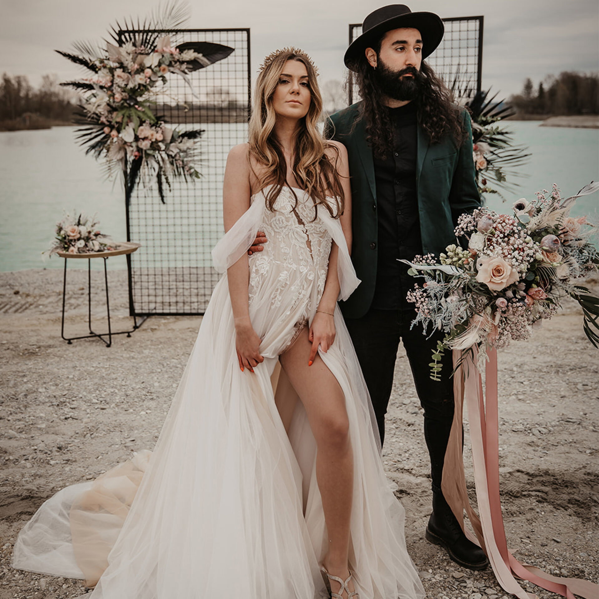 Das Brautpaar strahlt in einem stimmungsvollen Hochzeits-Setting am Wasser. Die Braut in einem romantischen Spitzenkleid und der Bräutigam im markanten Hut stehen vor einem Gitter, das mit Blumen verziert ist.