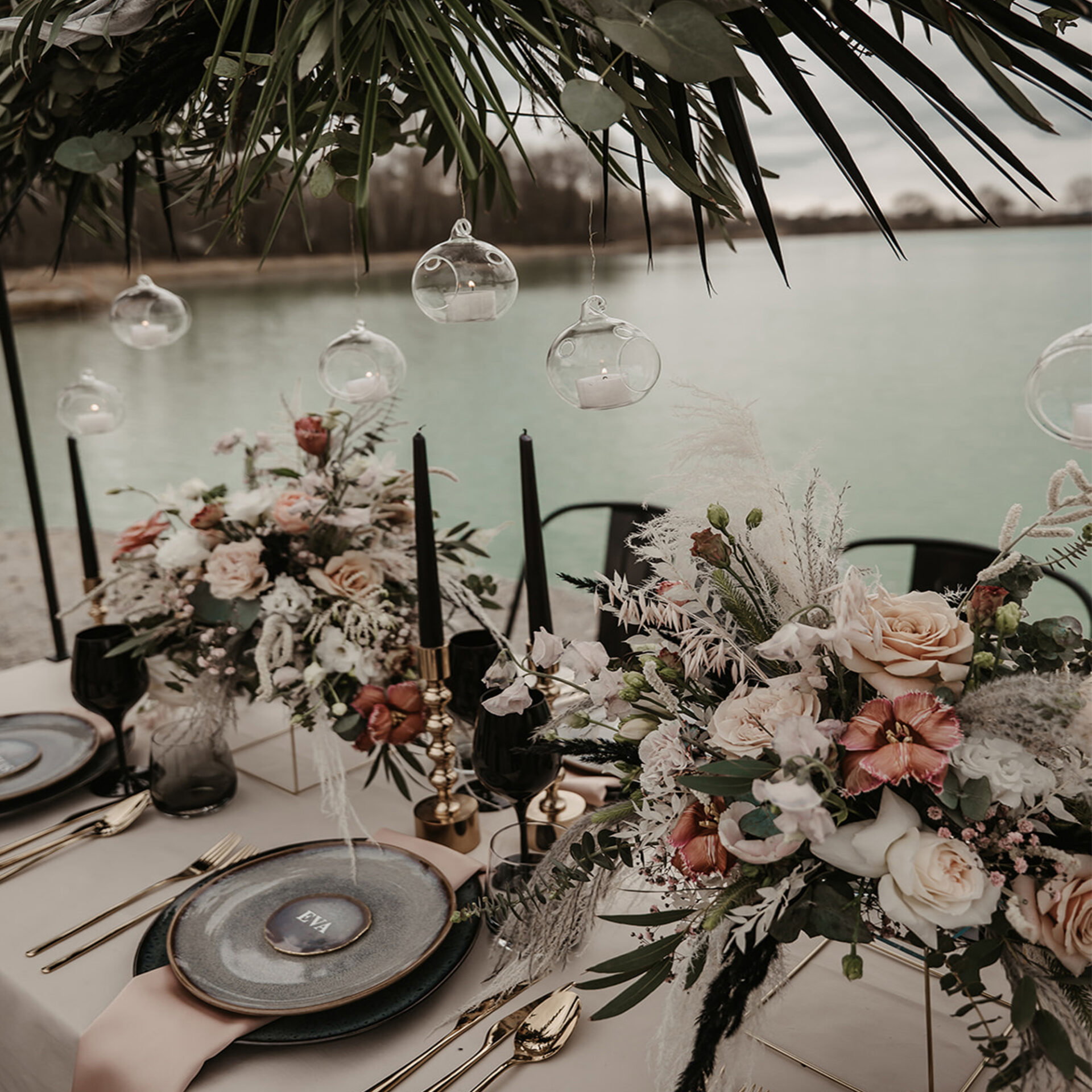 Hochzeitsdekoration mit floralen Gestecken und schwarzen Akzenten, eingerahmt von schwebenden Kerzen, die über einem festlich gedeckten Tisch am See hängen.