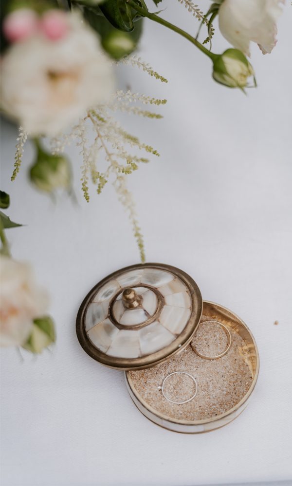 Ein stilvolles Ringset in einer goldenen Schmuckschatulle, platziert auf feinem Sand. Weiße Blüten im Hintergrund sorgen für eine sanfte, romantische Hochzeitsstimmung.