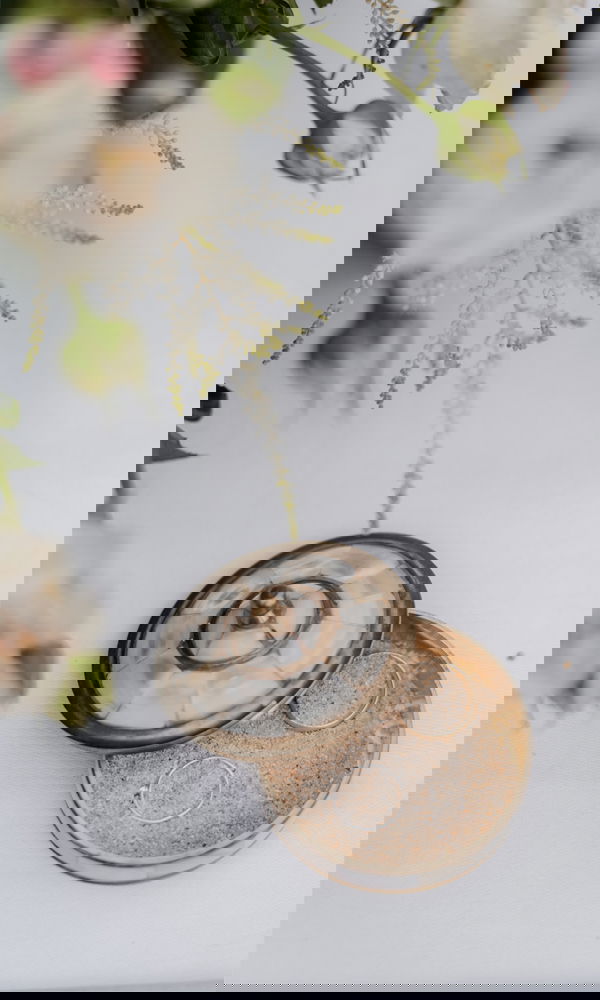 Ein stilvolles Ringset in einer goldenen Schmuckschatulle, platziert auf feinem Sand. Weiße Blüten im Hintergrund sorgen für eine sanfte, romantische Hochzeitsstimmung.