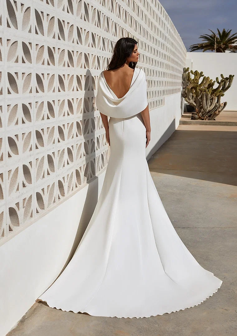Eine Braut trägt ein stilvolles, weißes Kleid mit tiefem Rückenausschnitt und bodenlanger Schleppe, die sanft über den Boden fällt, während sie an einer Wand mit geometrischen Formen vorbeigeht.