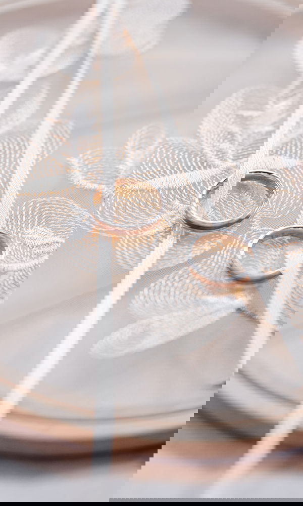 Zwei zarte Eheringe aus glänzendem Metall, stilvoll auf besticktem Stoff arrangiert. Feine Satinbänder verleihen der Inszenierung eine verspielte Leichtigkeit.