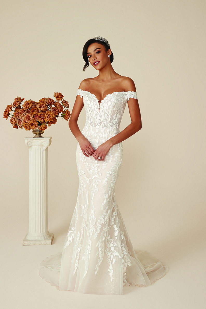 Modell in einem schulterfreien, figurbetonten Brautkleid mit filigranen Blumenapplikationen, steht in einem neutralen Raum neben einer klassischen Säule mit orangefarbenen Rosen.