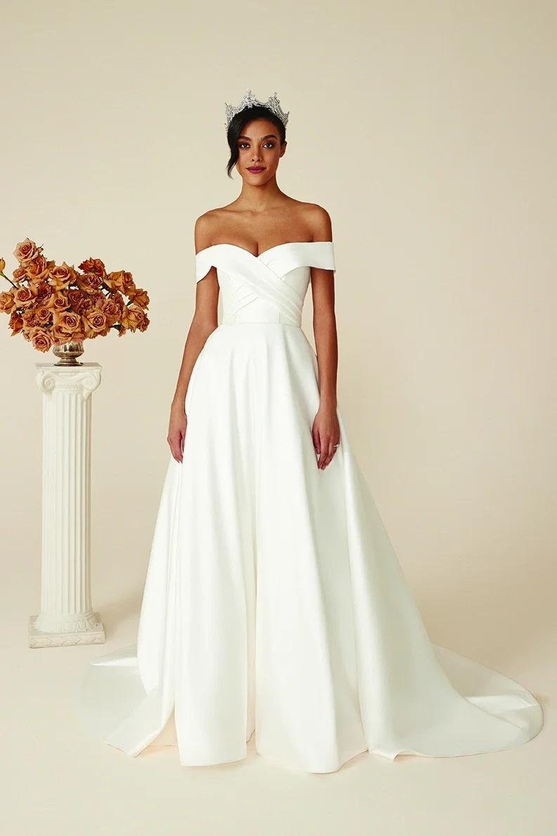 Eine Braut im schlichten, trägerlosen Ballkleid mit dramatischer Schleppe und Krone posiert in einem minimalistischen Raum, neben einer Säule mit rustikalen Blumen.