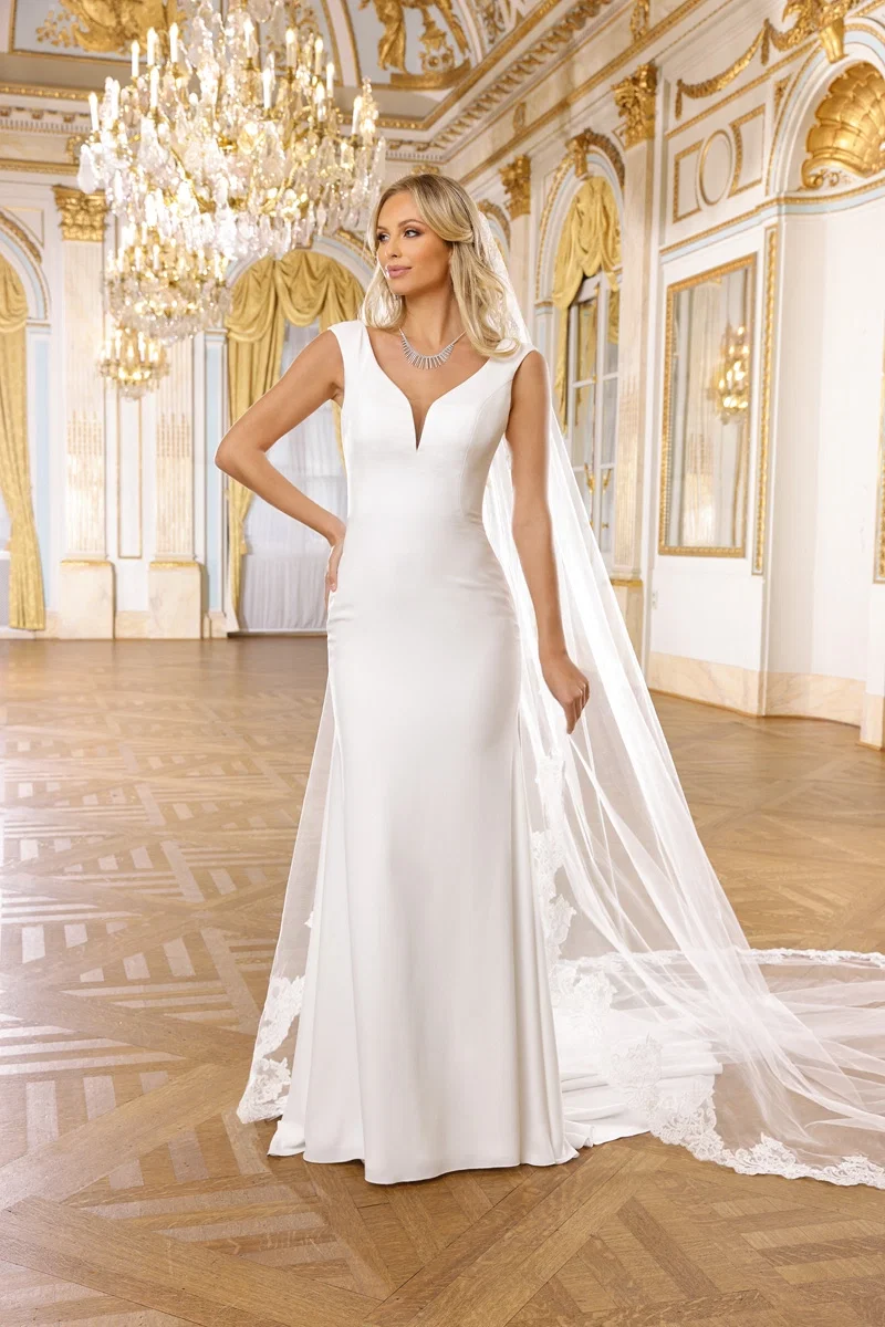Brautmodell in einem glatten, figurbetonten Brautkleid mit langer Schleppe und tiefem V-Ausschnitt, posiert in einem opulenten Ballsaal mit Kronleuchtern und kunstvoll dekorierten Wänden.