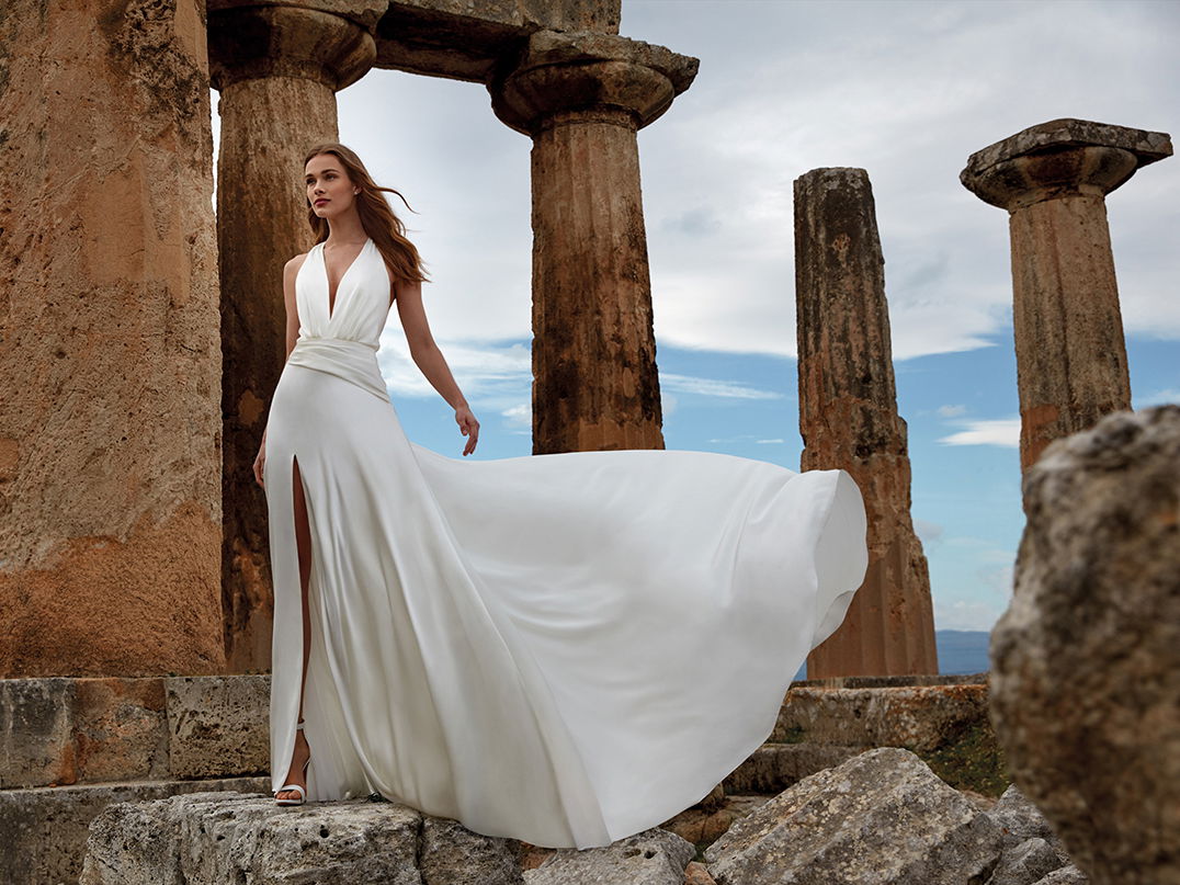 Modell in einem weißen, ärmellosen Brautkleid mit langer Schleppe und tiefem Beinschlitz, umgeben von antiken Ruinen und majestätischen Säulen.