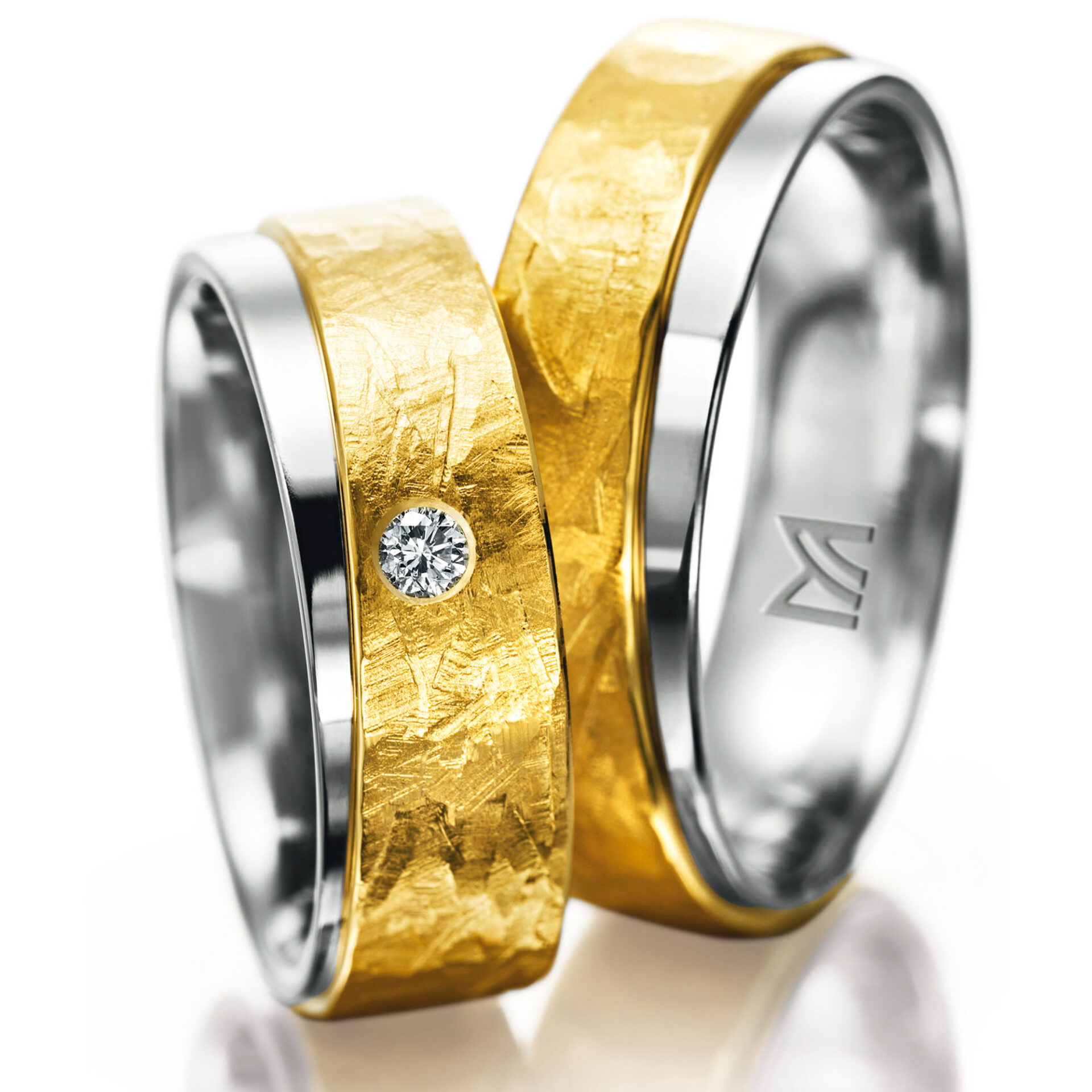 Zwei elegant gestaltete Eheringe, die eine Kombination aus glänzendem Weißgold und strukturiertem Gelbgold zeigen. Der breitere Ring ist mit einem Diamanten verziert, der in das Gelbgold eingelassen ist.