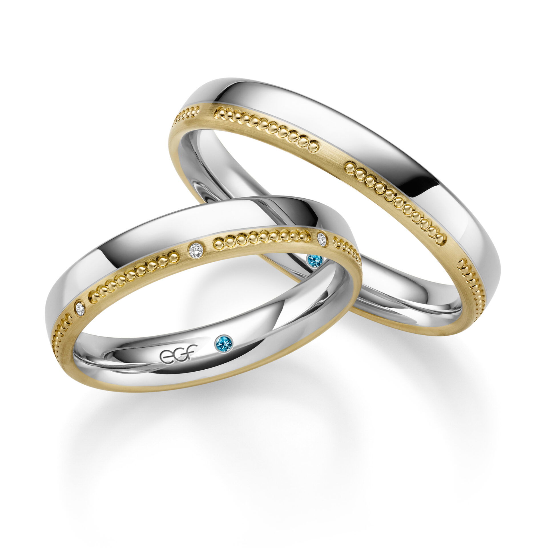 Ein Set aus Eheringen, die eine Kombination aus glänzendem Weißgold und satiniertem Gelbgold zeigen. Beide Ringe sind mit einer goldenen Kugelreihe und kleinen Diamanten, darunter ein blauer Edelstein, versehen.