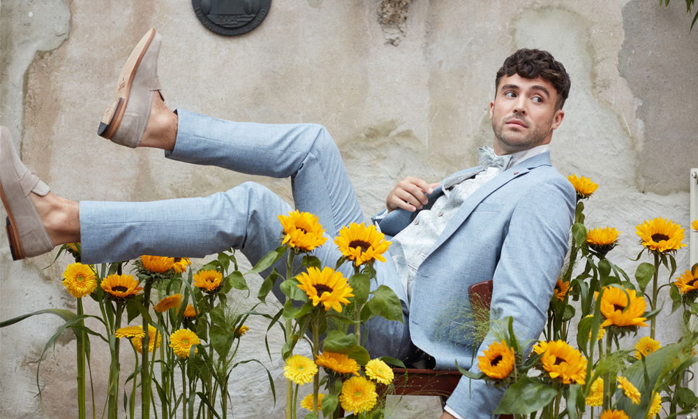 Ein junger Mann lehnt in einem hellen Anzug zurück, während er von einer Vielzahl strahlender Sonnenblumen umgeben ist. Im Hintergrund ist eine helle Wand zu sehen.