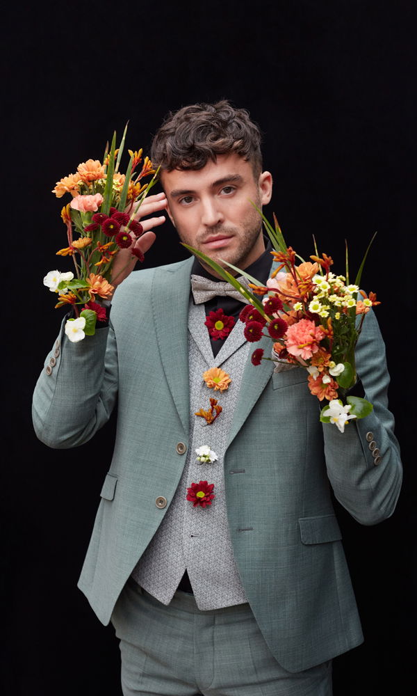 Porträt eines Mannes in einem grünen Anzug, dessen Look durch florale Elemente in Rot-, Orange- und Gelbtönen ergänzt wird, vor dunklem Hintergrund.